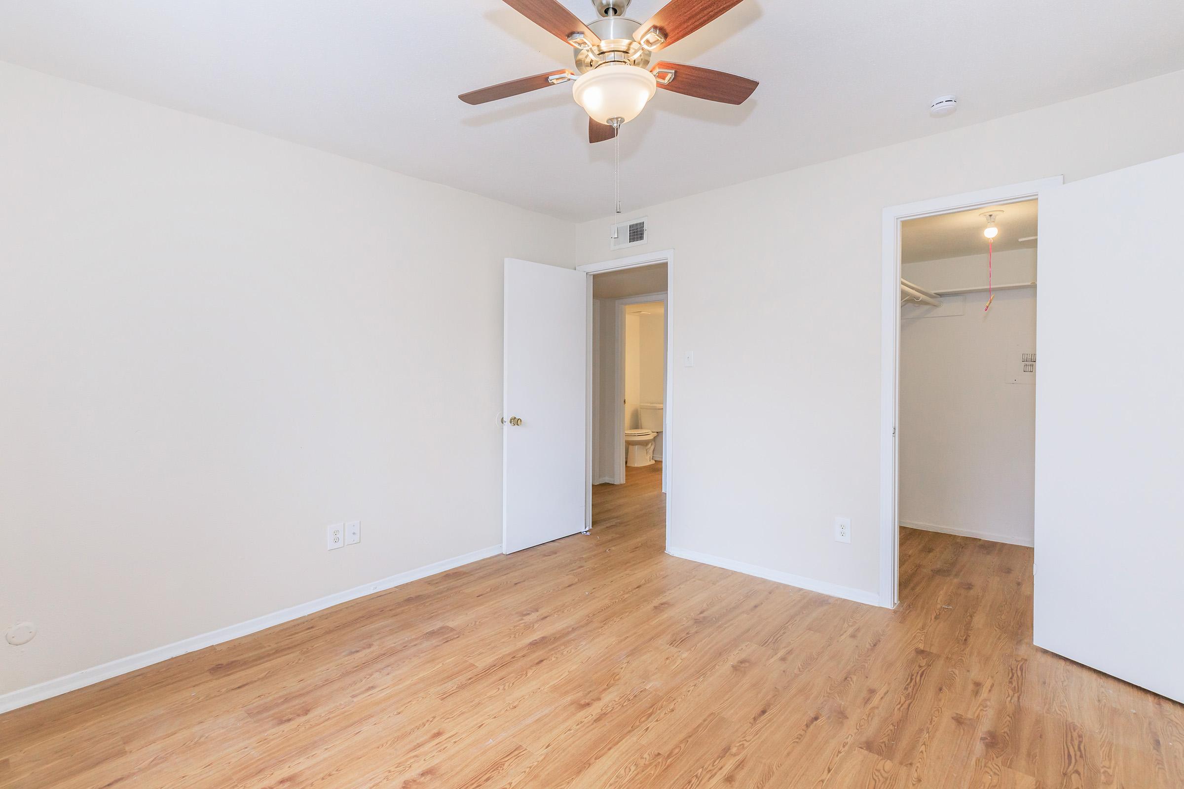 A bright, empty room featuring light wood flooring, a ceiling fan with a light fixture, and a doorway leading to a bathroom. Opposite, there is another open door leading to a closet area. The walls are painted in a soft beige color, giving a spacious and airy feel.