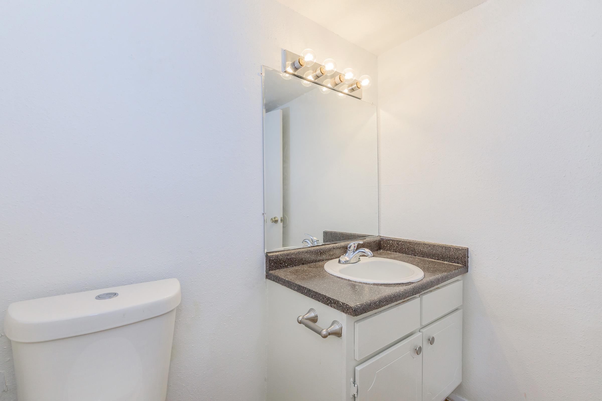 A simple bathroom featuring a white toilet, a small vanity with a sink, and a wall-mounted mirror above the sink. The walls are painted white, and there are light fixtures above the mirror providing illumination. The vanity has a dark countertop and white cabinetry.