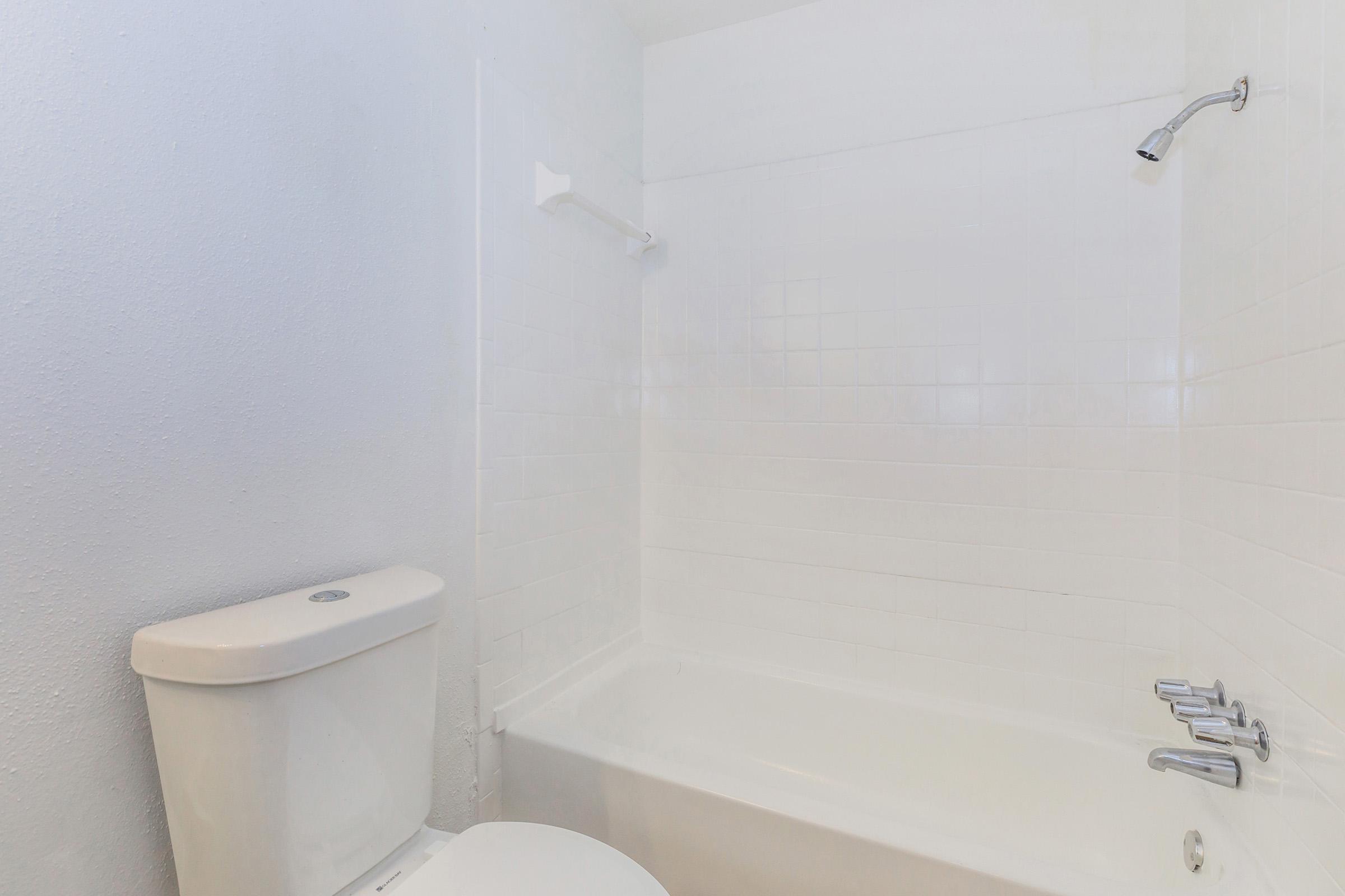 A clean, minimalist bathroom featuring a white bathtub, a toilet, and a showerhead. The walls are tiled in a light color, contributing to a bright and spacious appearance. The setting is uncluttered, emphasizing simplicity and functionality.