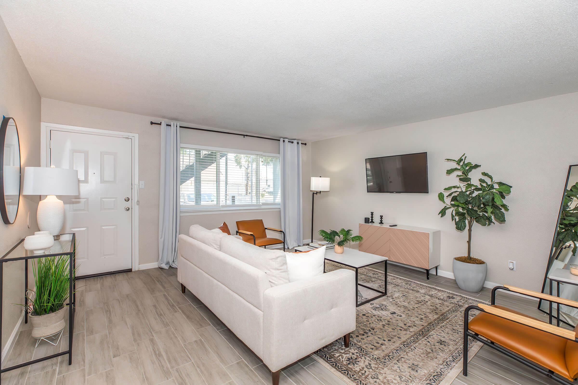 A cozy living room featuring a light-colored sofa, two stylish chairs, a glass coffee table, and a decorative rug. Natural light streams in through a window with sheer curtains. A modern TV is mounted on the wall opposite the sofa, and a tall indoor plant adds a touch of greenery to the space.