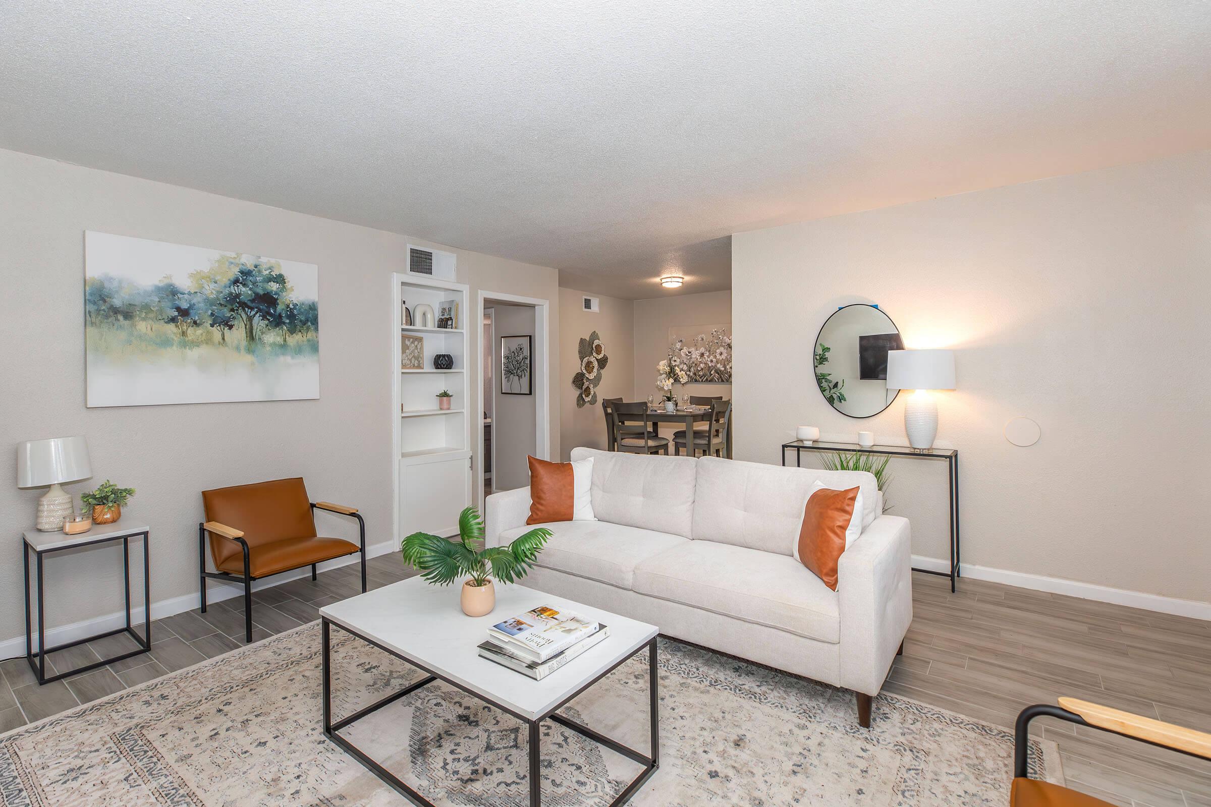 Cozy living room with a light-colored sofa adorned with orange pillows, a coffee table with magazines, and green plants. A stylish armchair is in one corner. The background features a dining area with a modern table, decorative wall art, and a round mirror that reflects soft lighting.