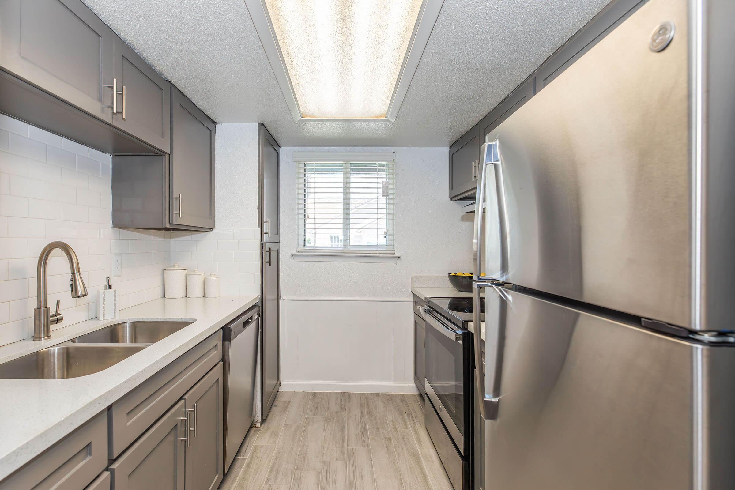 Modern kitchen featuring stainless steel appliances, including a refrigerator and stove. The gray cabinets complement the light-colored countertops. A window allows natural light to enter, and a sink is positioned between the counter space. Stylish tile backsplashes enhance the contemporary design.