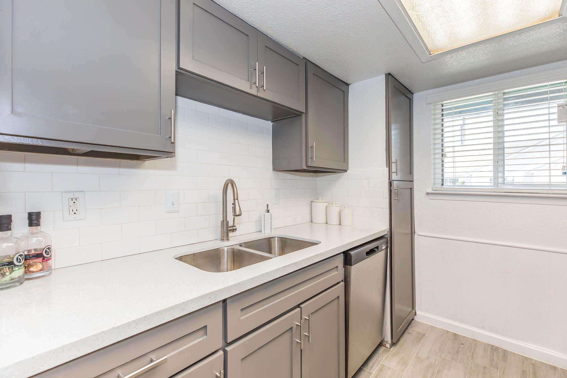 Modern kitchen featuring gray cabinetry, a double sink, and stainless steel appliances. Light-colored countertop and a tiled backsplash complement the space. A window lets in natural light, and decorative storage containers are placed on the countertop.