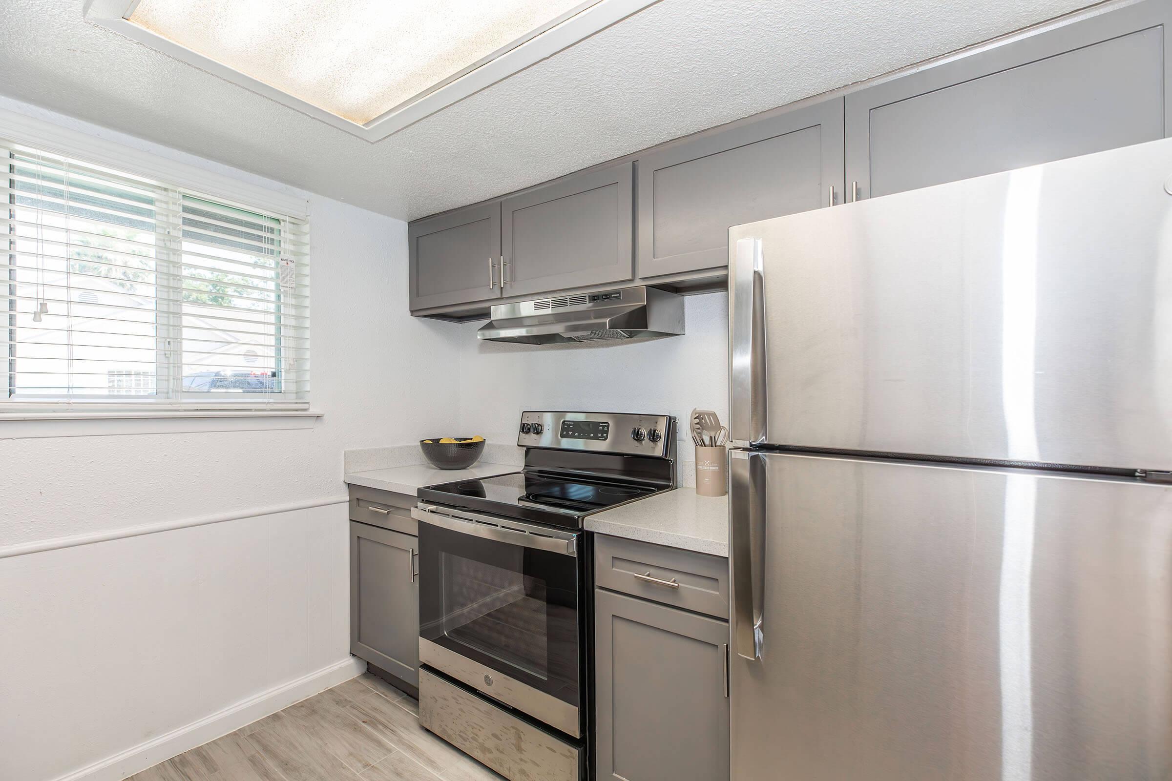 Modern kitchen featuring stainless steel appliances including a stove, refrigerator, and microwave. Grey cabinetry and light countertops create a sleek look. There's a window with blinds allowing natural light, and a bowl of fruit on the countertop adds a welcoming touch.