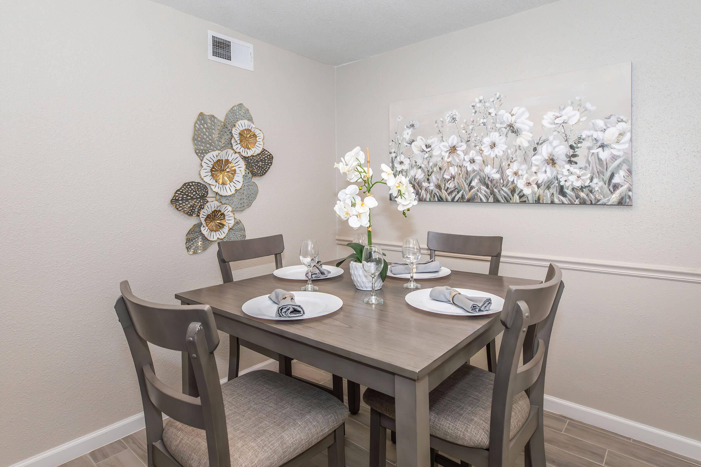 A cozy dining area featuring a light-colored wall with a large floral painting, a wooden table set for four with plates and napkins, and a decorative vase with white orchids. The chairs are simple and modern, creating an inviting atmosphere.