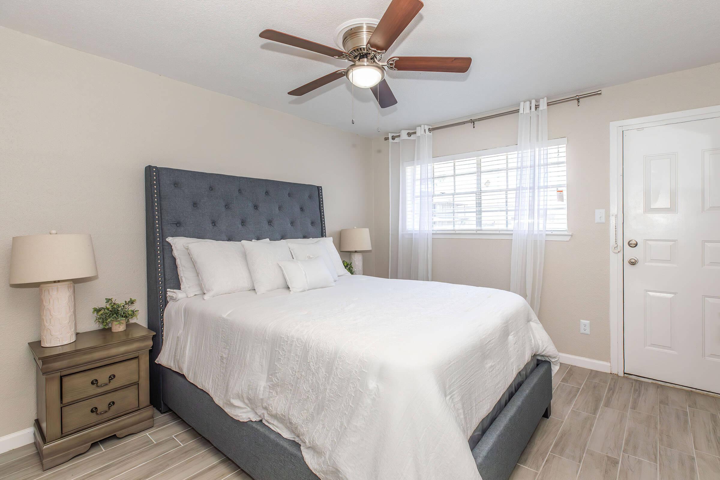 A cozy bedroom featuring a blue upholstered bed with white bedding, two bedside lamps, and a wooden nightstand. A ceiling fan hangs above, and a window with sheer curtains allows natural light to brighten the space. The walls are painted a soft beige, creating a warm and inviting atmosphere.