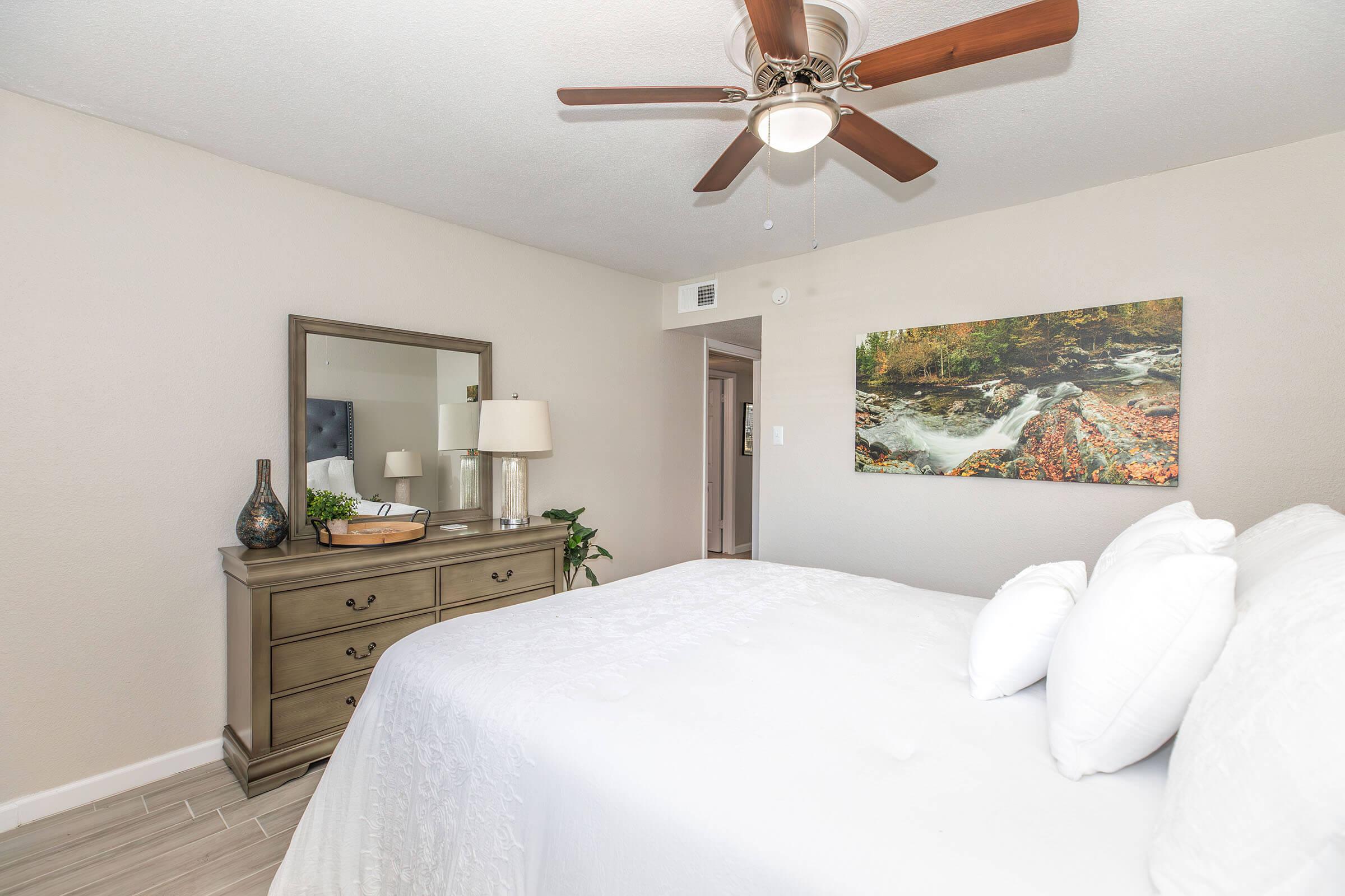 A comfortable bedroom featuring a queen-sized bed with white linens, a wooden dresser with a lamp and decor items, and a mirror. A nature-themed painting hangs on the wall, and there’s a ceiling fan above. The walls are light-colored, creating a cozy atmosphere.