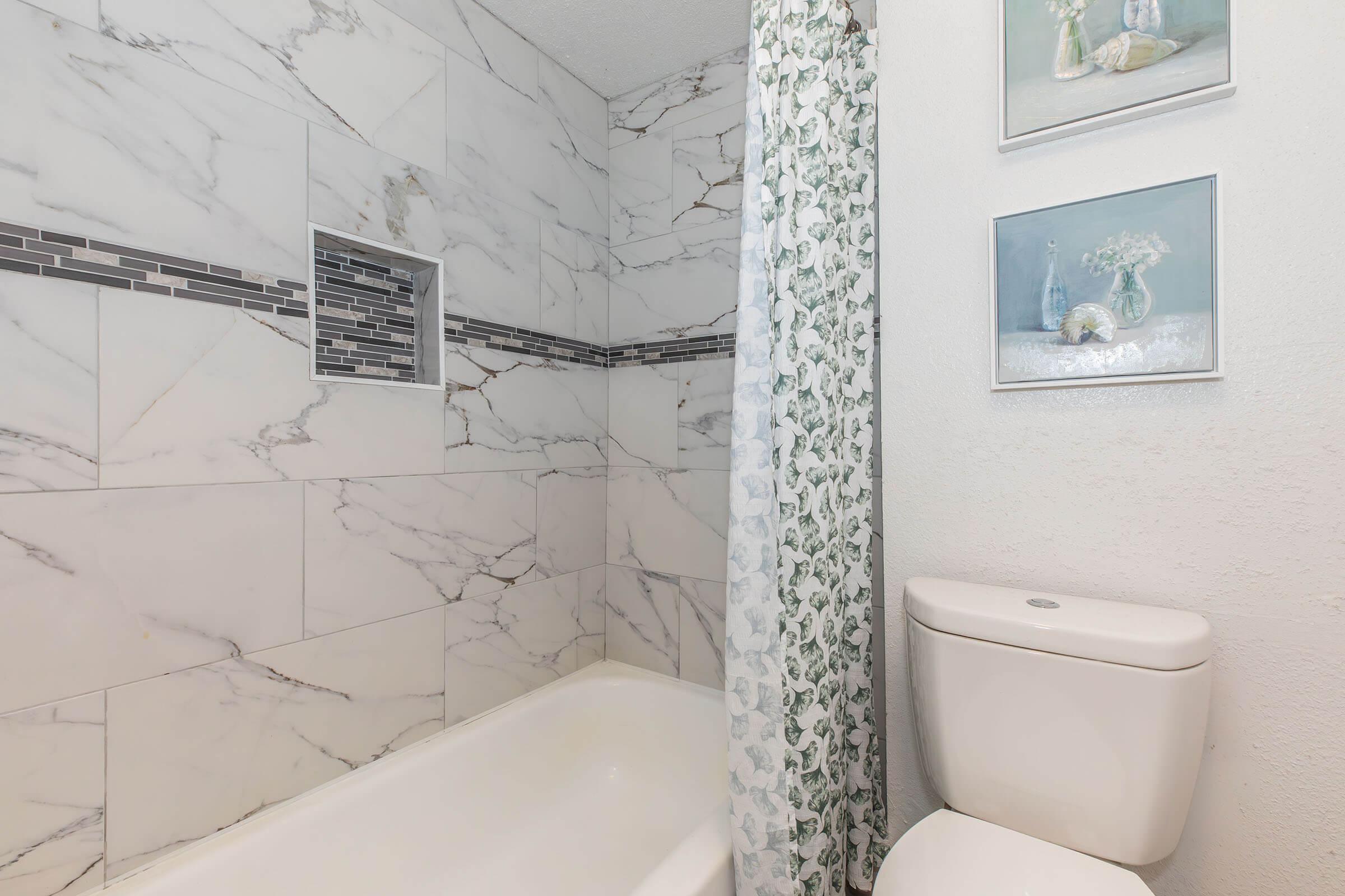 Modern bathroom featuring a white tub, a white toilet, and marble wall tiles with a decorative stripe. A floral shower curtain adds a touch of color, and two framed pictures hang on the wall, enhancing the decor. Natural light brightens the space, creating a clean and inviting atmosphere.