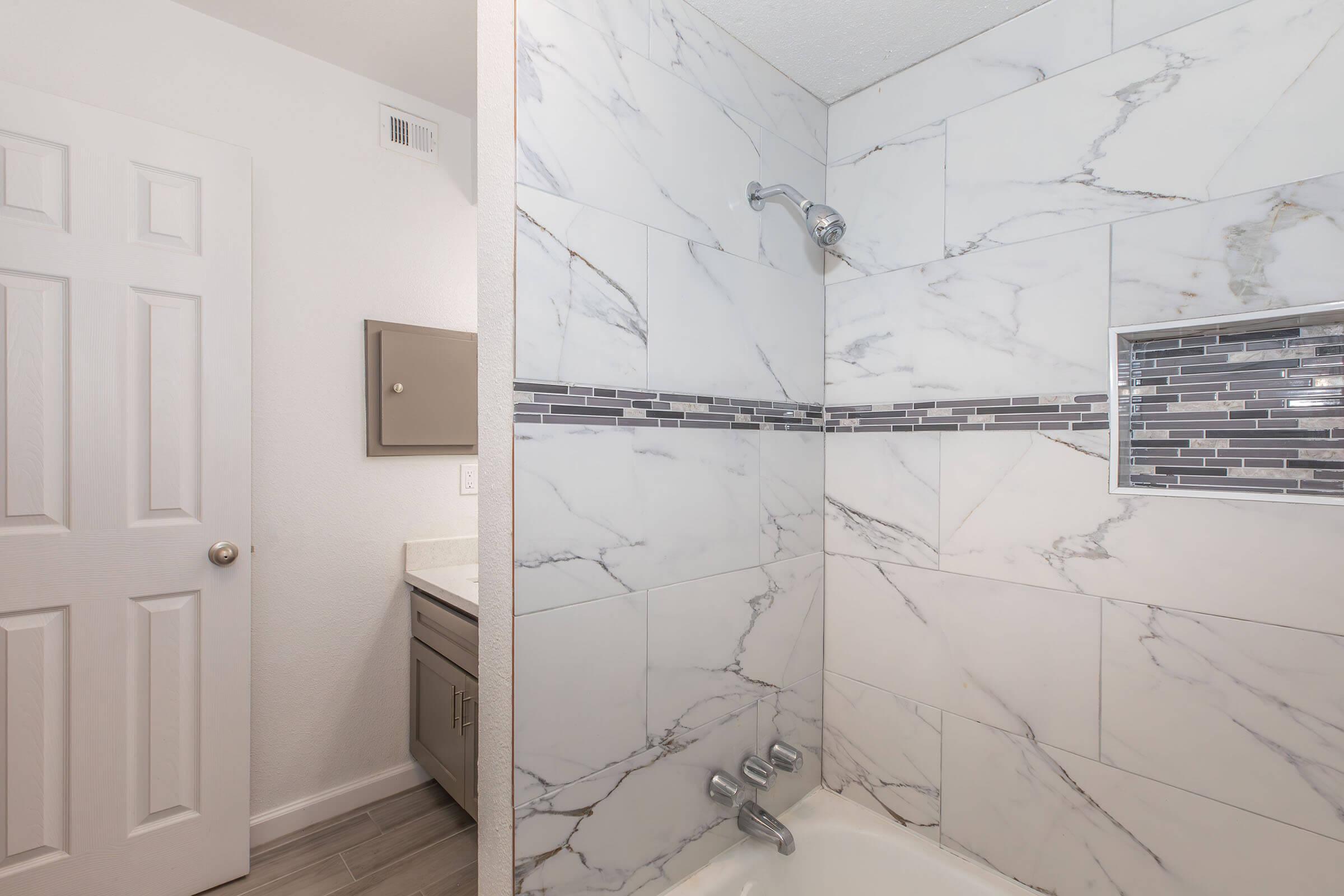 A modern bathroom featuring a shower with marble tile walls, a built-in shelf with decorative tiles, and a sleek faucet. The room is bright with natural light, and a door leads to another space. A vanity with a mirror is also visible, giving a clean and contemporary look.