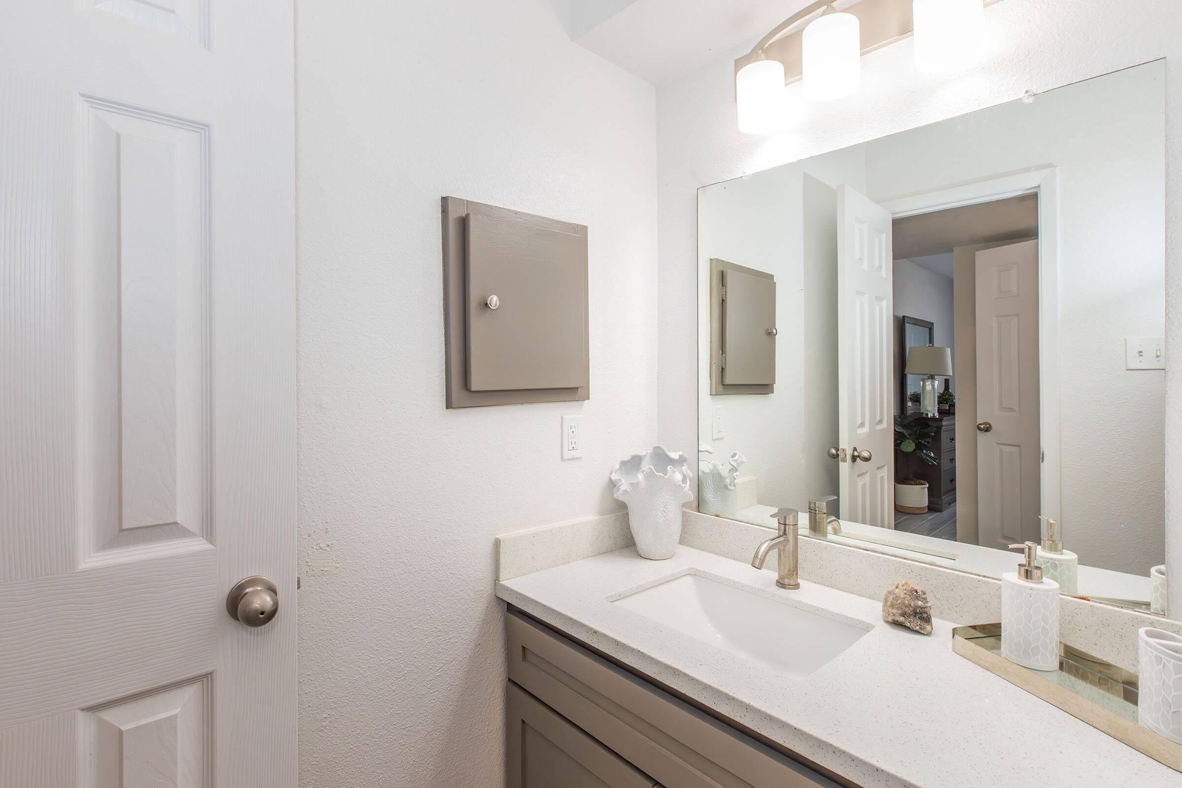 A clean and bright bathroom featuring a modern sink with a faucet, a large mirror above the sink, a light fixture, and a small decorative plant. A cabinet and door are visible, adding to the room's functional design. The walls are painted white, contributing to a fresh and airy atmosphere.