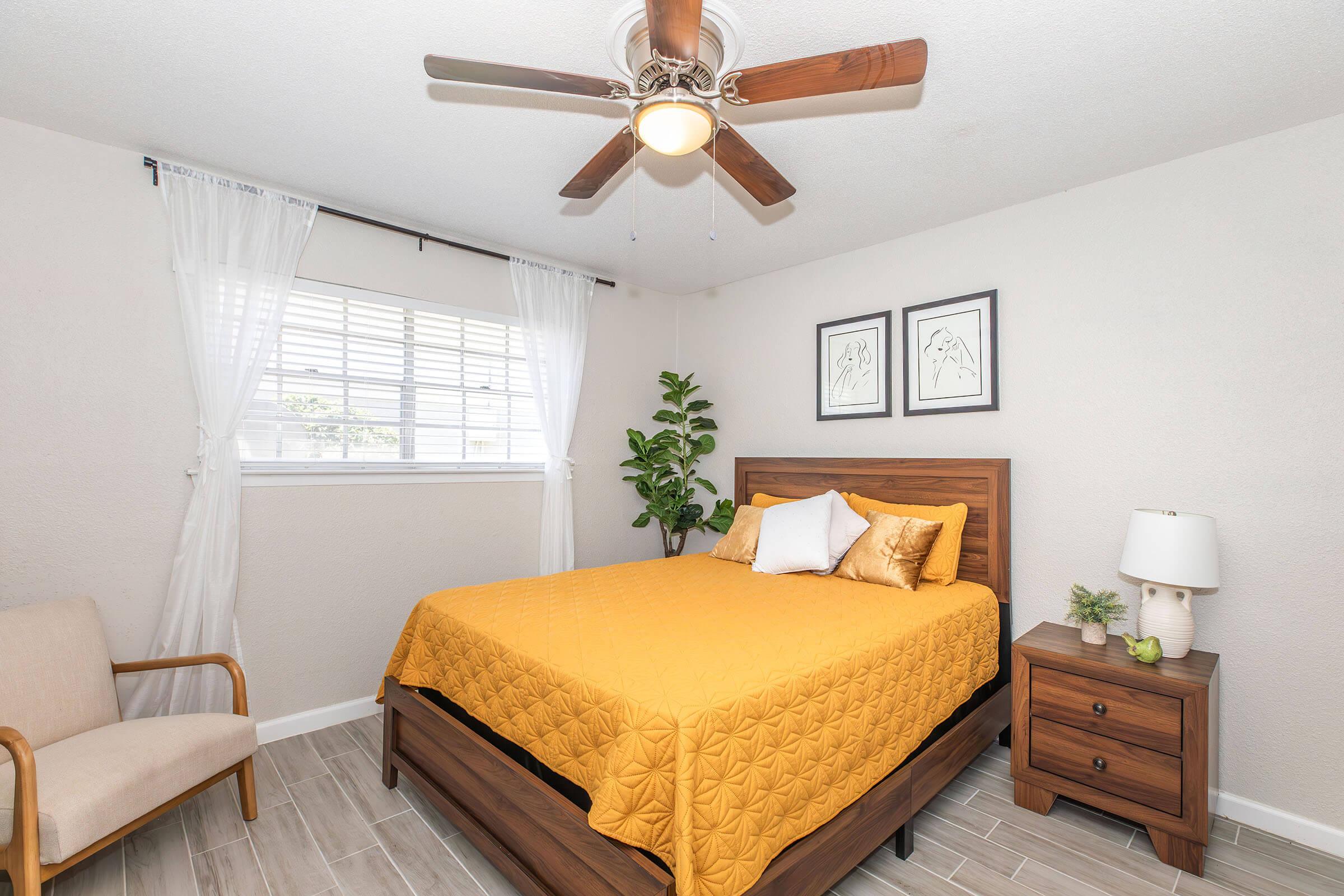 A well-lit bedroom featuring a queen-sized bed with an orange quilt, two decorative pillows, and a wooden headboard. There's a light-colored chair in the corner, a small bedside table with a lamp and a plant, and framed artwork on the wall. Sheer white curtains adorn the window.