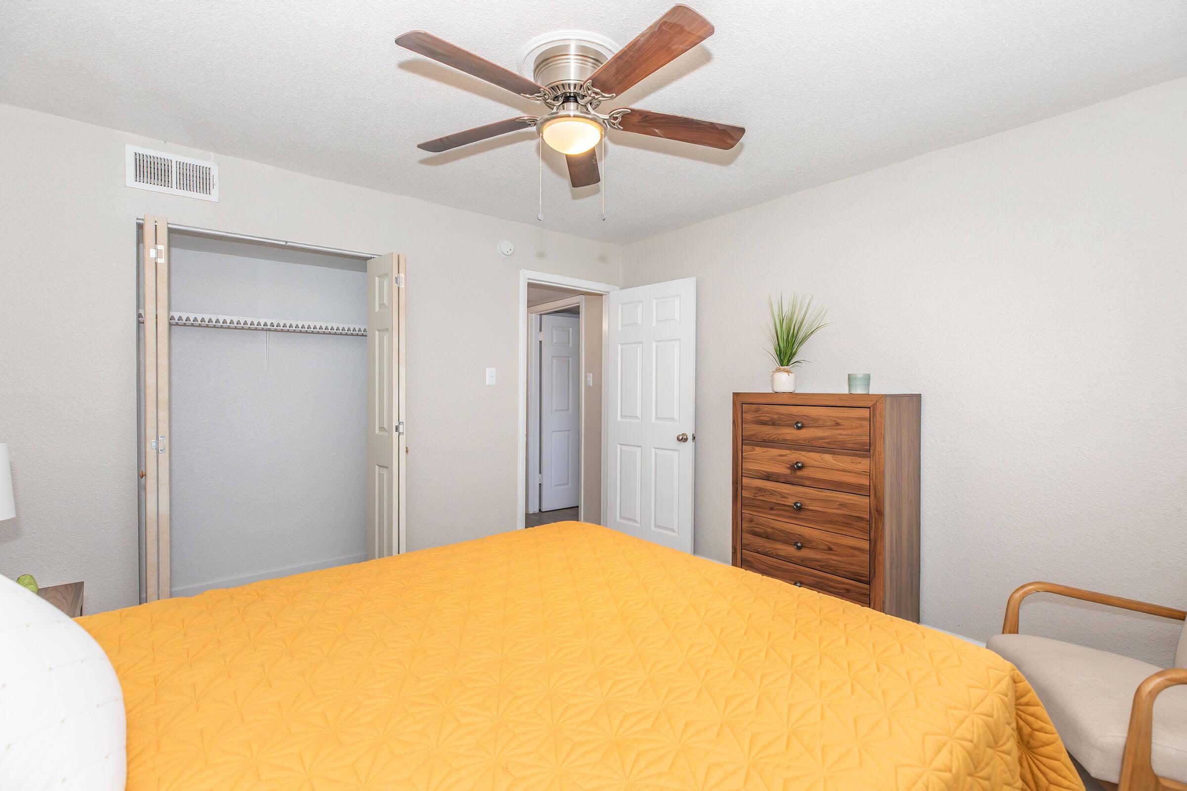 A cozy bedroom featuring a queen-sized bed with a bright yellow quilt, a wooden dresser, and a comfortable chair. A ceiling fan is visible, and there’s a door leading to a closet on the left. The walls are painted in a soft beige color, creating a warm and inviting atmosphere.