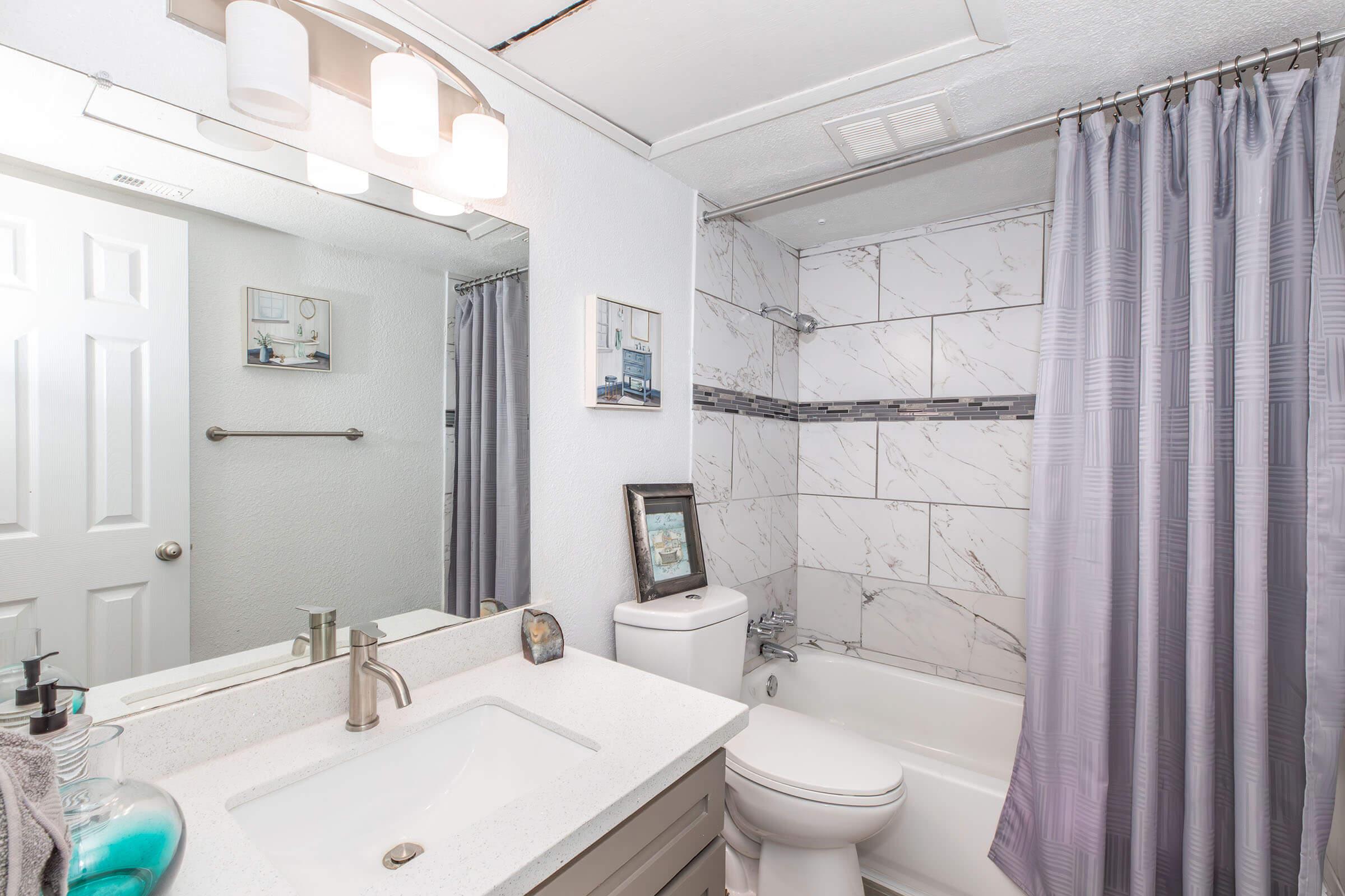 A modern bathroom featuring a white marble-themed shower area, a clean white sink with a sleek faucet, and a large mirror. Soft gray curtains hang by the bathtub, and wall decor adds a touch of style. The overall design is bright and contemporary, creating an inviting atmosphere.