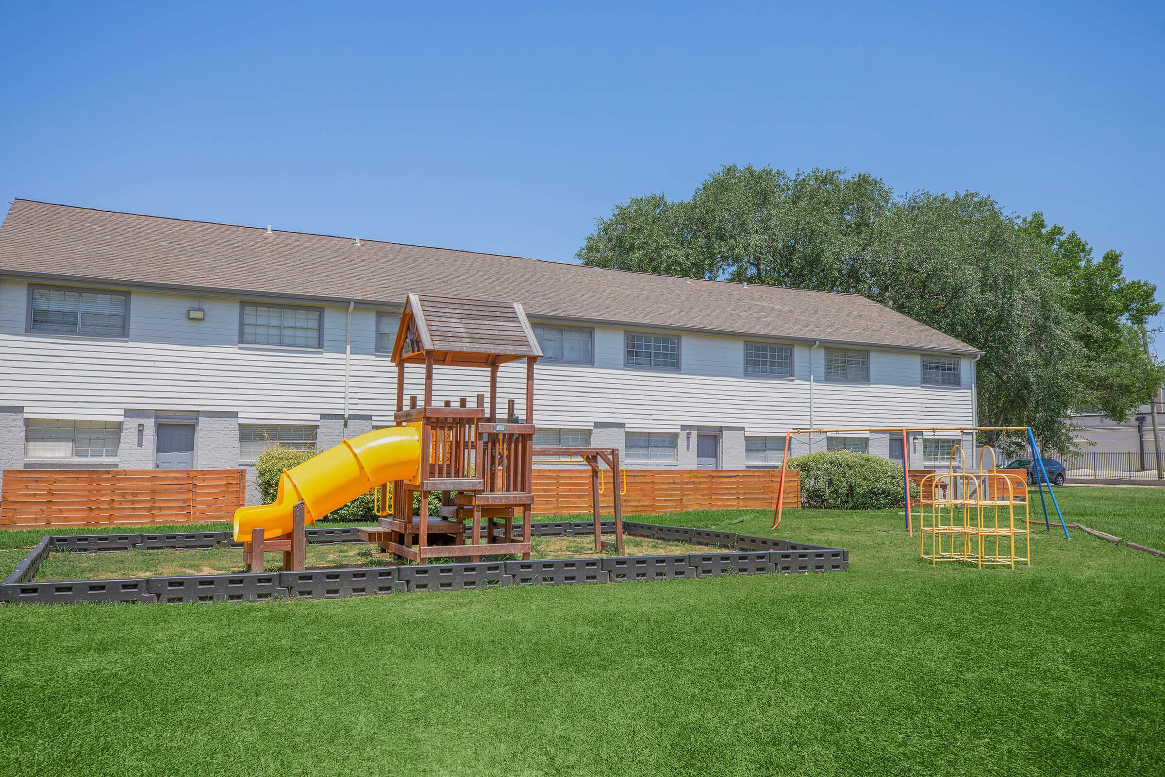 A playground featuring a wooden play structure with a yellow slide, surrounded by a black border. Nearby, there are swings on a metal frame and a grassy area. In the background, there are several two-story residential building units under a clear blue sky.