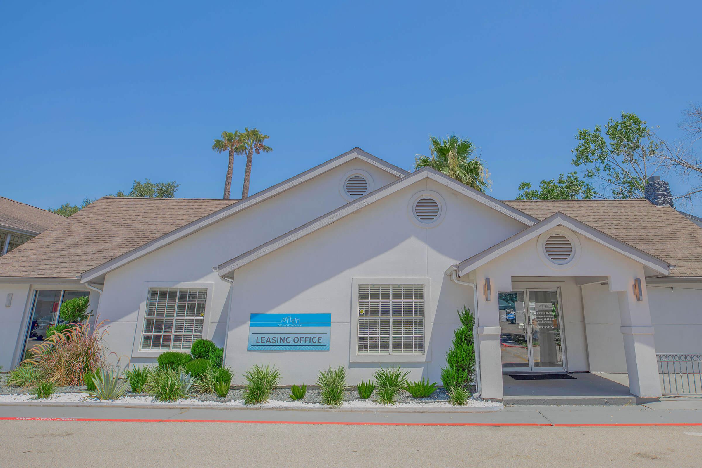 A well-lit leasing office building with a sloped roof, featuring large windows and a glass entrance door. The sign on the front indicates "Leasing Office." Surrounding the entrance are landscaped plants and small palms, set against a clear blue sky.