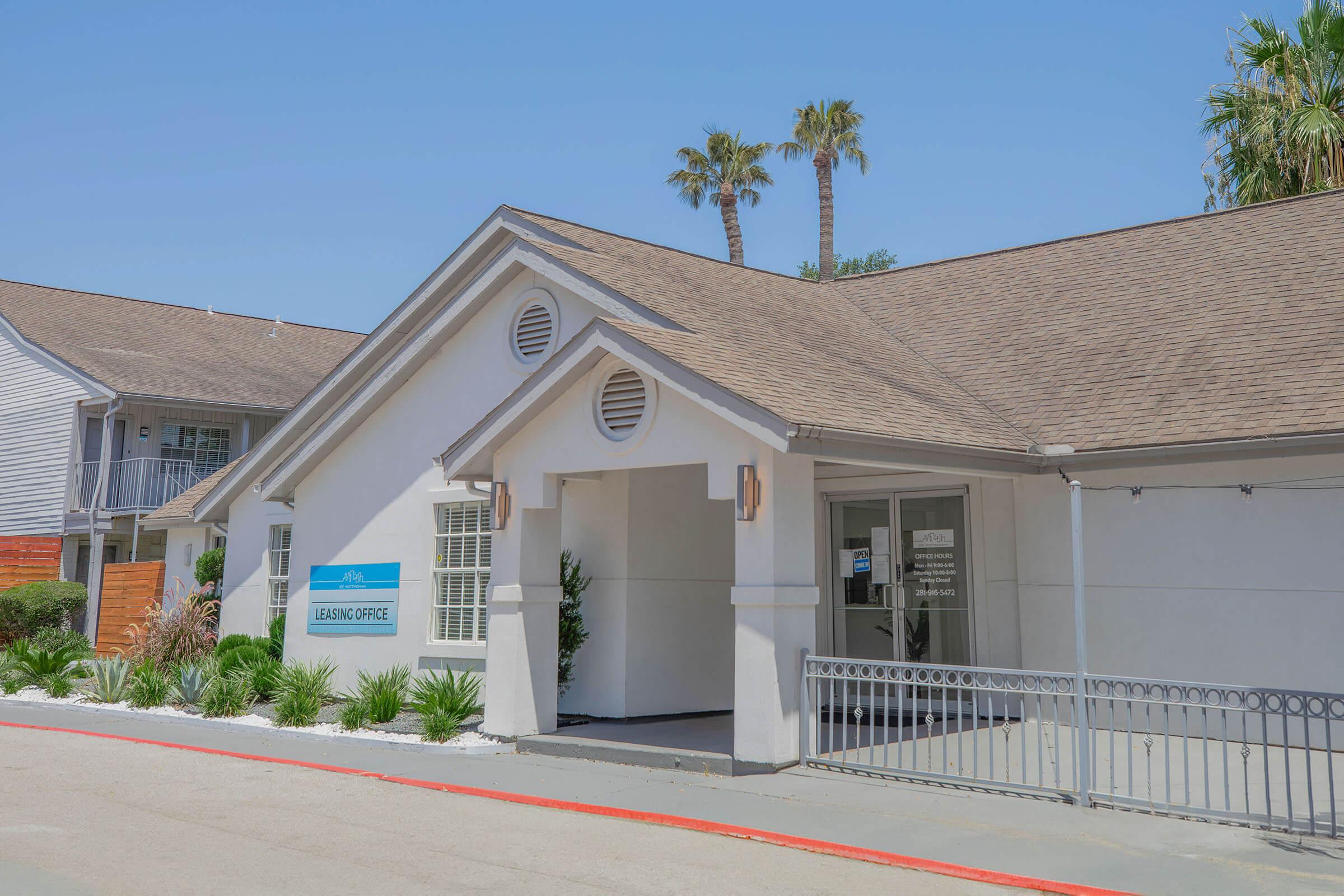A leasing office building with a white exterior and a peaked roof, featuring a sign that reads "Leasing Office." The entrance is framed by two windows, and there are palm trees in the background against a clear blue sky. The area is landscaped with greenery and a stone pathway.