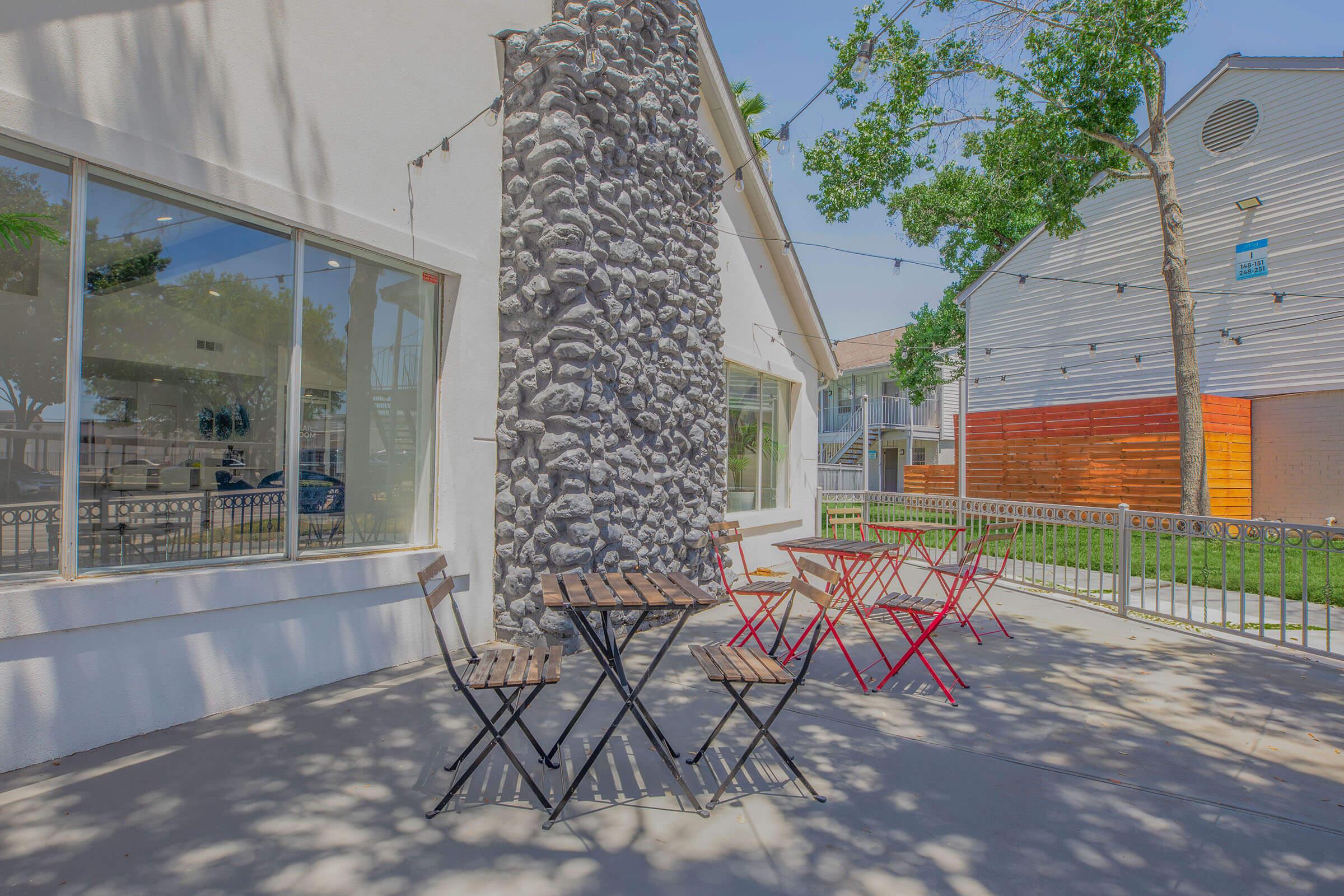A quiet outdoor seating area featuring a stone column, small wooden tables, and colorful chairs. Sunlight brightens the space, surrounded by trees and a building in the background. The scene invites relaxation and conversation in a casual setting.