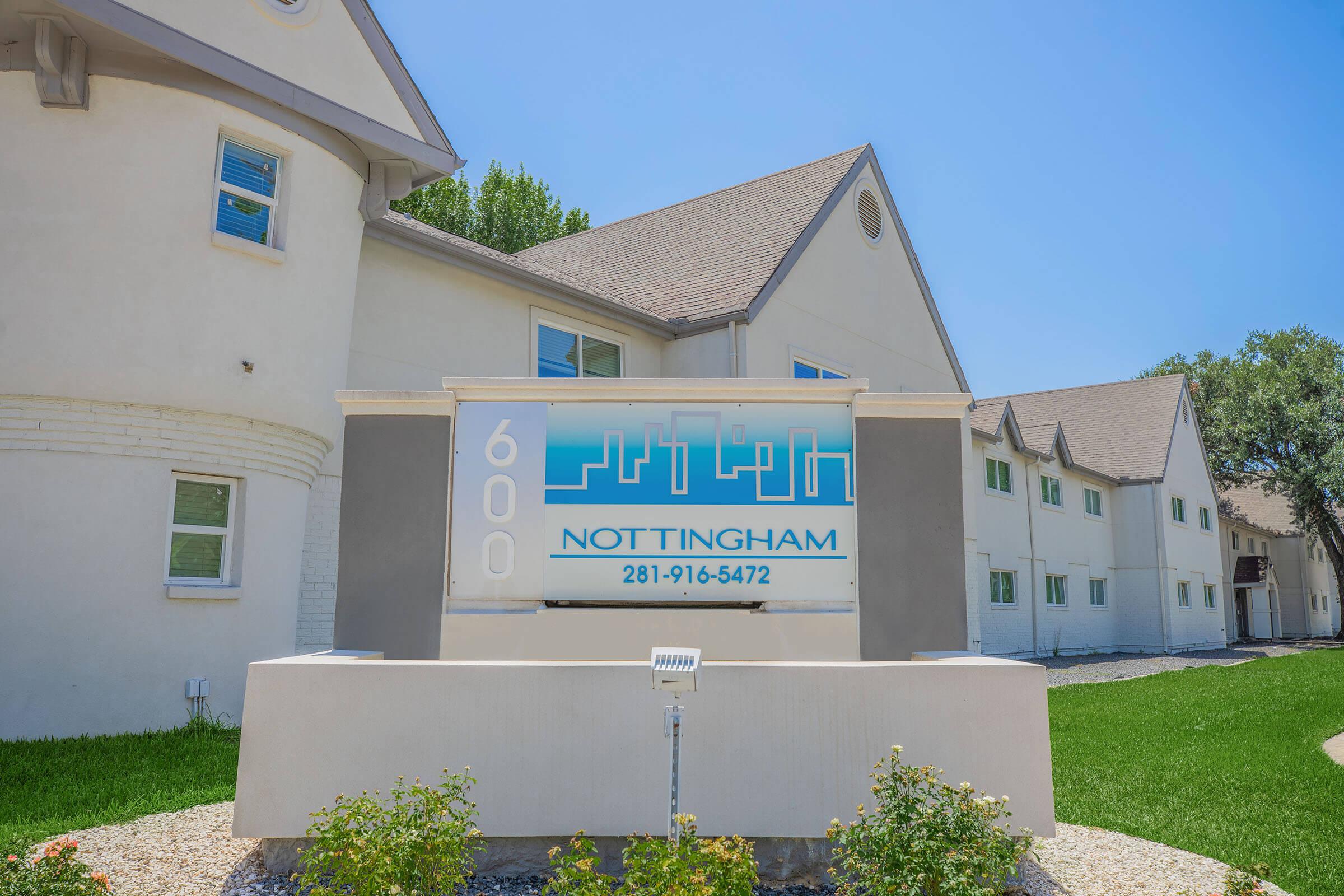 Sign for Nottingham, located at 600, displaying the phone number 281-916-5472. The sign is set in front of a residential building with a modern architectural design, surrounded by green grass and shrubs under a clear blue sky.