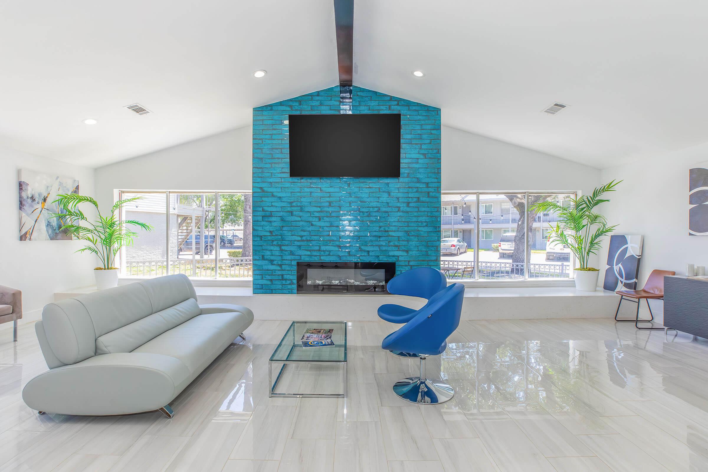 Bright and modern living room featuring a turquoise accent wall with a mounted TV, a white curved sofa, a blue chair, and large windows allowing natural light. There's a minimalist coffee table and a sleek side cabinet, along with decorative plants enhancing the contemporary aesthetic.