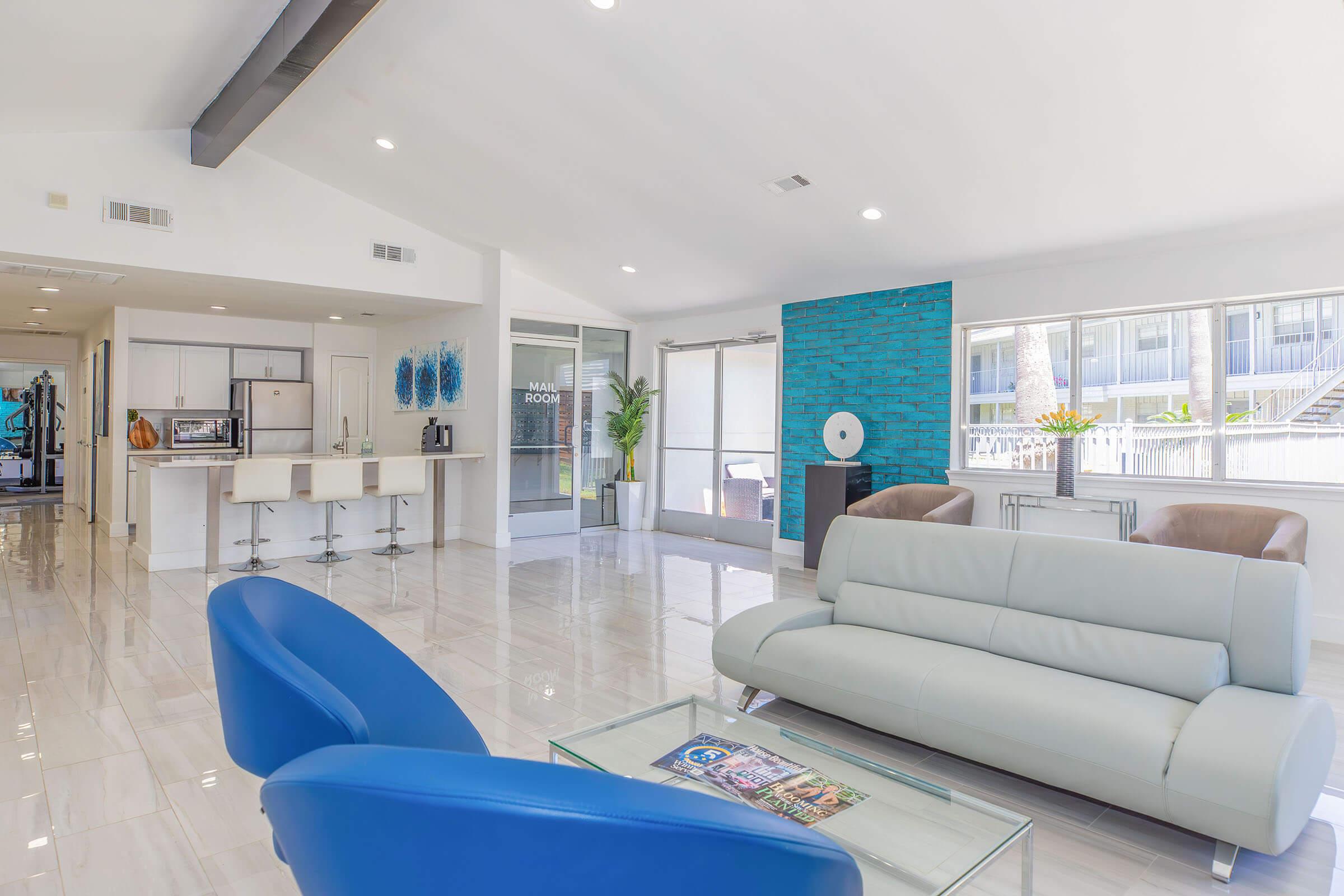 A modern, bright living space featuring a light-colored sofa and blue accent chairs. The room has large windows, providing natural light, and includes a contemporary kitchen area in the background. There's a coffee table in the center and stylish decor on the walls, creating an inviting atmosphere.