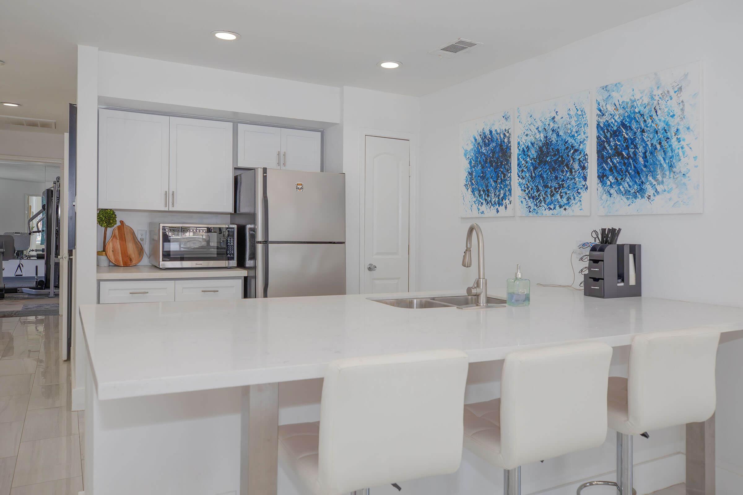 Modern kitchen with white cabinetry and a large island featuring a sink. Stainless steel appliances, including a refrigerator and microwave, are visible. The space is decorated with abstract blue artwork on the wall and features three white bar stools. Bright, clean layout with ample lighting.