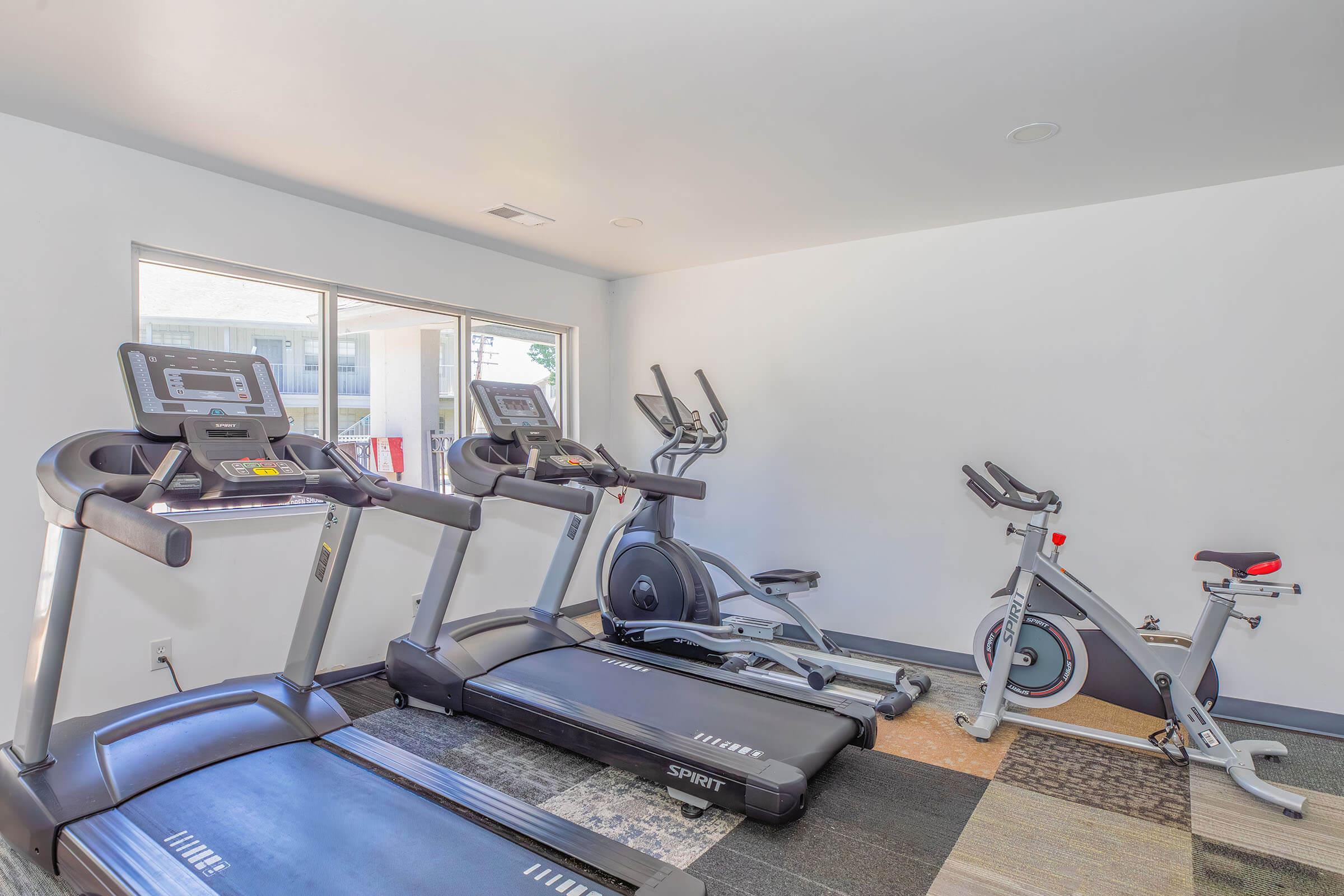 A fitness room featuring three exercise machines: two treadmills, an elliptical machine, and a stationary bike. The space is bright with natural light coming from a window and has a modern design with various flooring textures.