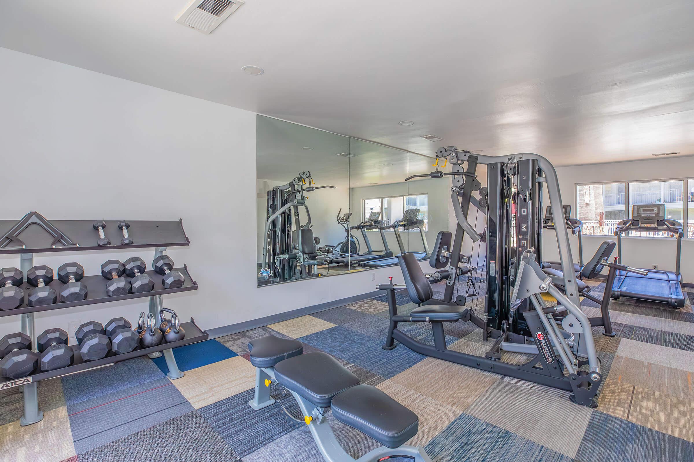A modern gym interior featuring a variety of exercise equipment. Visible items include a weight rack with dumbbells, a multi-station workout machine, a bench for exercises, and cardio machines like a treadmill. The space is well-lit with a large mirror reflecting the equipment. Stylish flooring adds to the aesthetic.