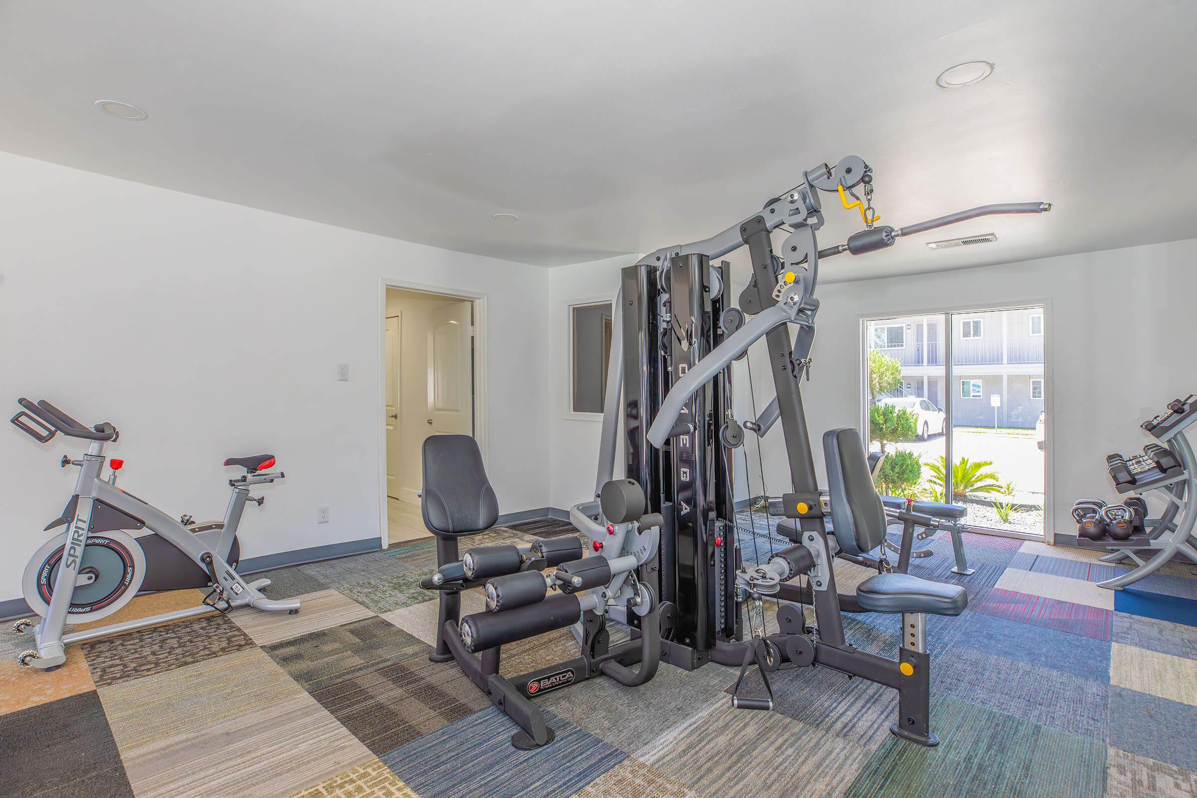 A well-equipped gym space featuring a multi-station workout machine, a stationary bike, and free weights. The room has a modern design with colorful carpet tiles and plenty of natural light coming from windows.