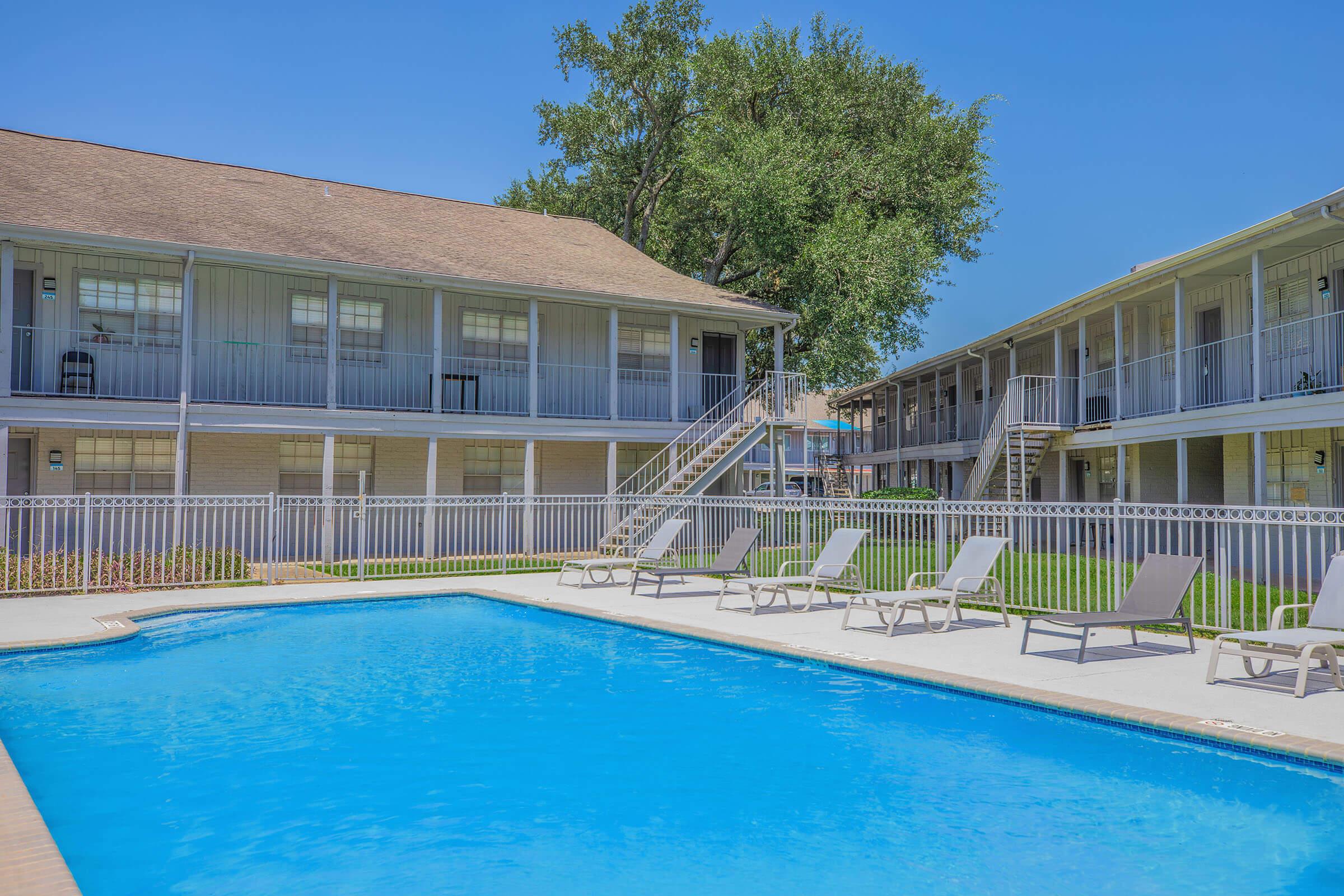 A crystal-clear swimming pool surrounded by lounge chairs, with two multistory buildings in the background. The setting features well-maintained grass and trees, under a bright blue sky. Stairs lead to upper levels of the buildings, creating a welcoming atmosphere for relaxation.