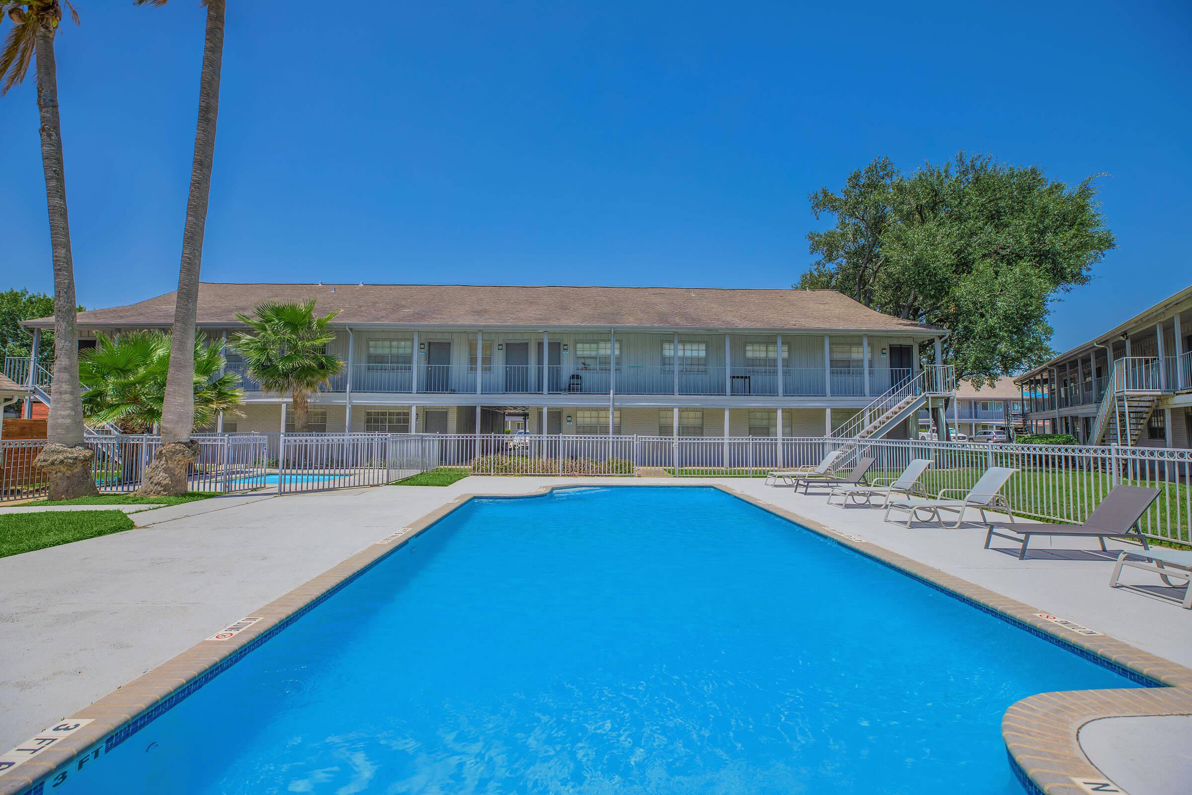 A clear blue swimming pool surrounded by lounge chairs, with a two-story building in the background featuring balconies. Palm trees and greenery line the pool area, set against a bright blue sky.