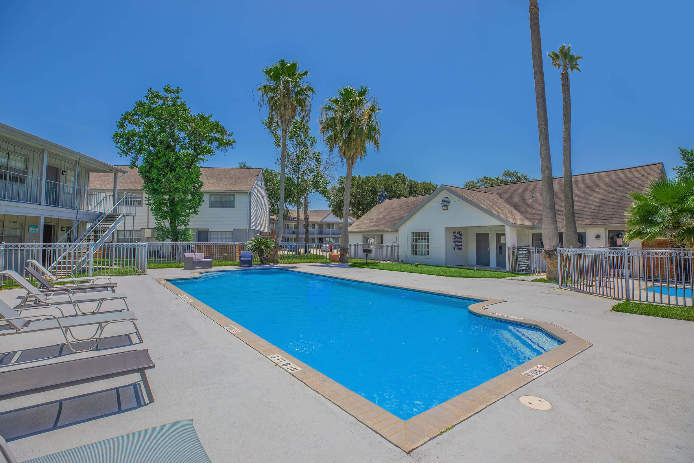 A clear blue swimming pool surrounded by lounge chairs and palm trees, with residential buildings in the background. The scene is bright and sunny, showcasing a tranquil outdoor setting.