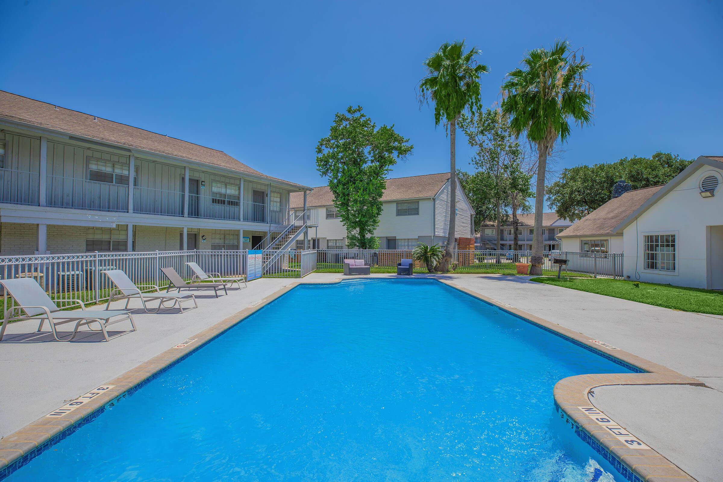 A bright sunny day at an outdoor swimming pool surrounded by lounge chairs. The pool is clear blue, with a well-maintained lawn and palm trees nearby. In the background, there are several apartment buildings featuring balconies. The atmosphere is inviting and perfect for relaxation.