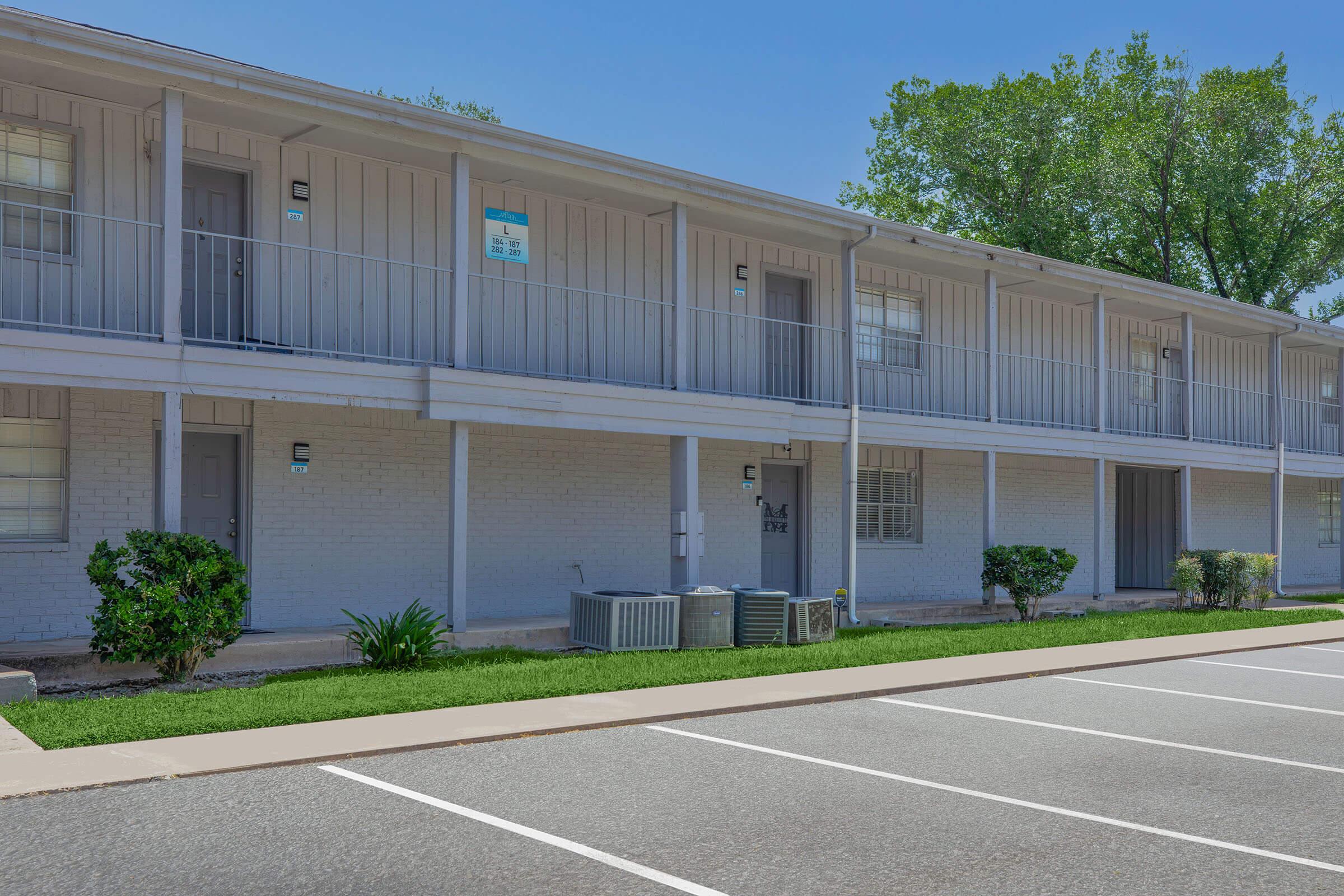 Two-story residential building with a row of apartments. The exterior features light-colored siding and small balconies. Below, there are air conditioning units, neatly trimmed shrubs, and a paved parking lot with marked spaces. The sky is clear and blue, suggesting a bright day.