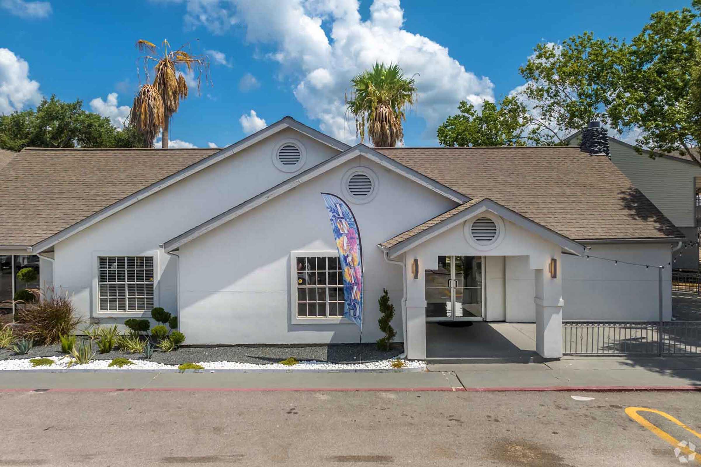 A single-story white building with a sloped roof, featuring large front windows. A colorful banner is displayed near the entrance, which is framed by decorative plants. The background includes a clear blue sky with fluffy clouds and palm trees. A paved pathway leads up to the building's entrance.
