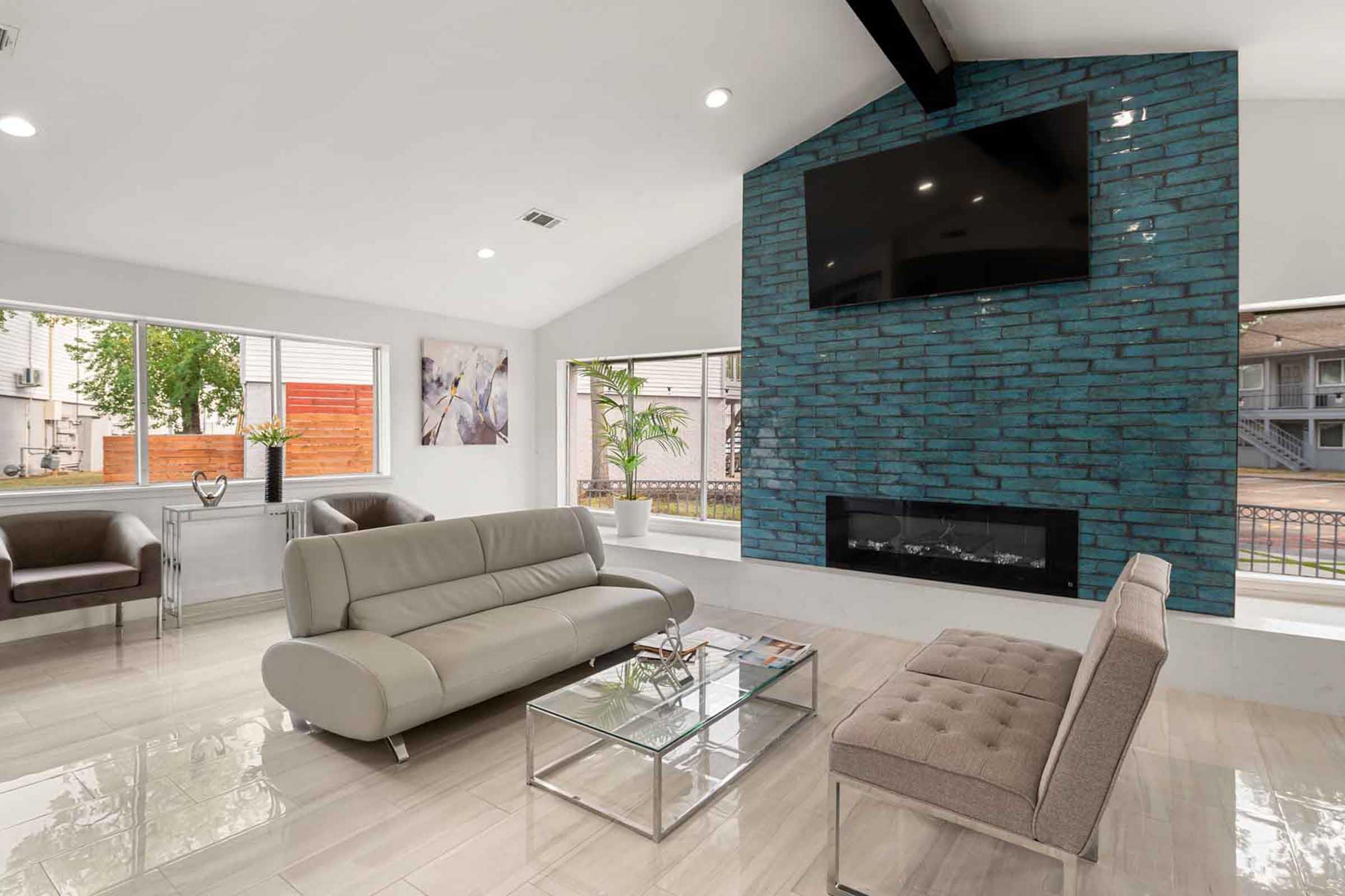 A modern living room featuring a light-colored sofa and a beige bench, with a stylish glass coffee table. The room has large windows allowing natural light, showcasing a blue brick feature wall with a mounted TV above a sleek fireplace. Decor includes indoor plants and contemporary furnishings.