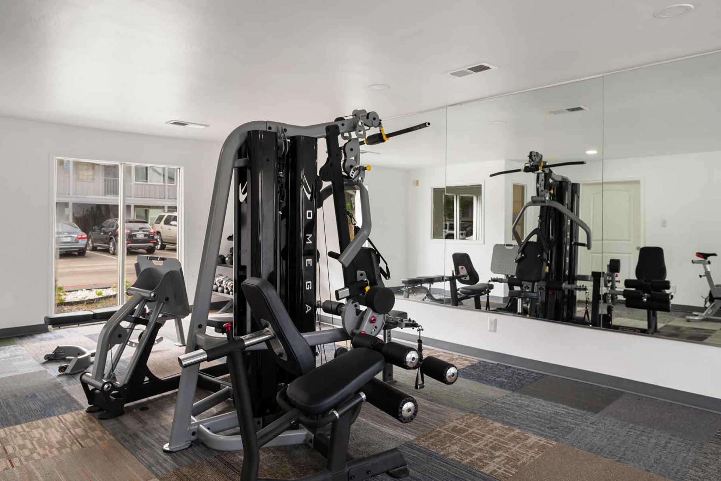 A modern gym interior featuring various exercise equipment, including a multi-station weight machine, a bench, and cardio machines. Mirrors line one wall, reflecting the equipment and enhancing the sense of space in the brightly lit room with neutral colors and carpeted flooring.