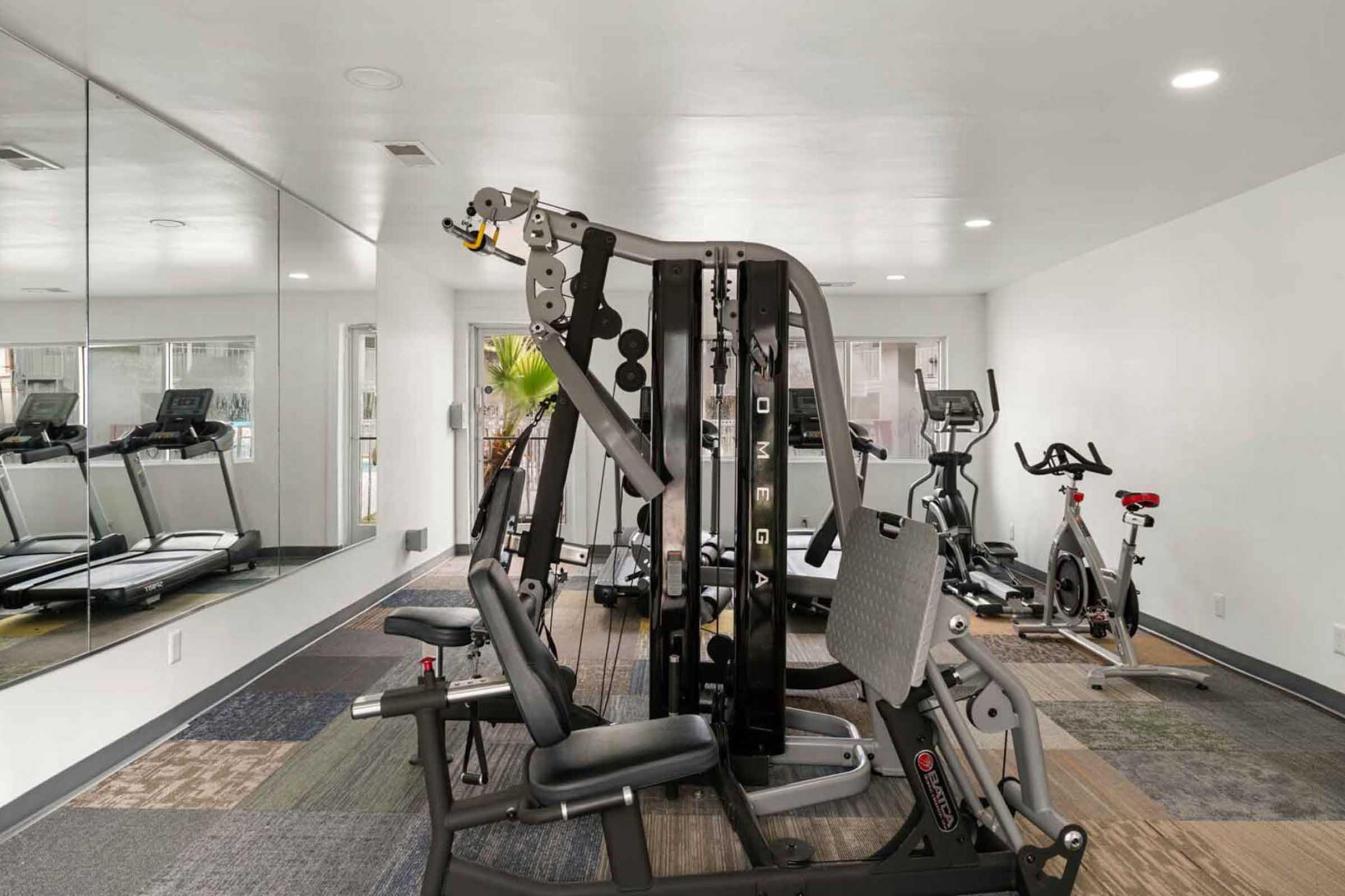 A modern gym interior featuring a multi-station weight machine, treadmills, and stationary bikes. The space is well-lit with mirrors on one wall and includes various workout equipment on a carpeted floor, creating a functional and inviting fitness environment.