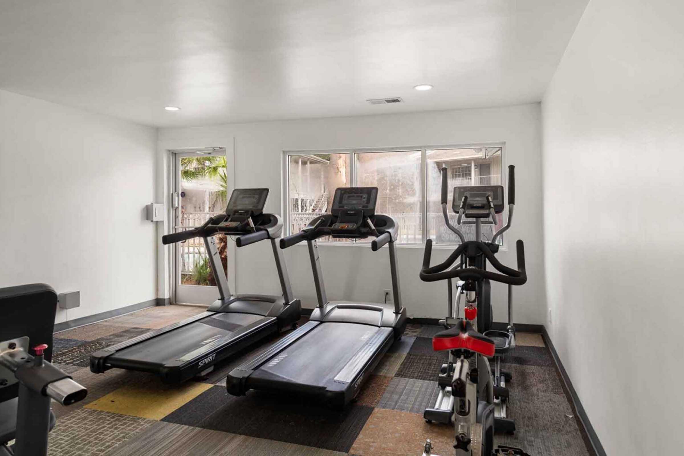 Interior of a gym featuring two treadmills and an exercise bike. The space is well-lit with large windows providing natural light. The flooring has a patterned design, and there is a door leading outside. The gym appears clean and organized, suitable for workout sessions.