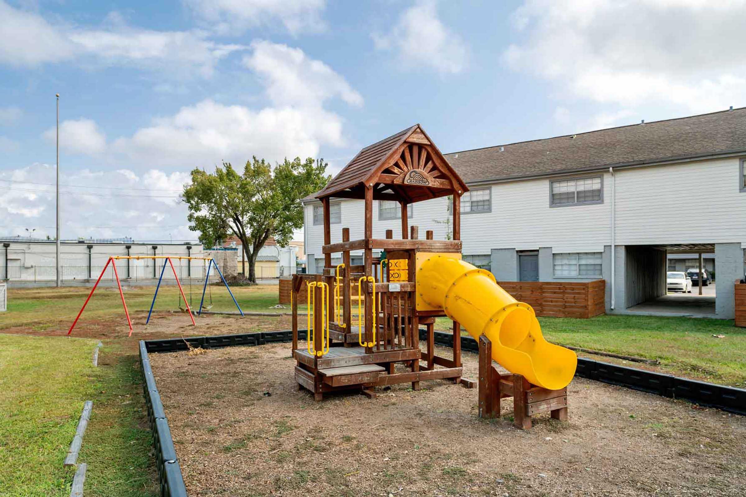 A wooden playground structure featuring a yellow slide and climbing area, surrounded by an open grassy space. In the background, there are swings attached to a frame and a series of residential buildings. The sky is partly cloudy, and the area appears well-maintained.