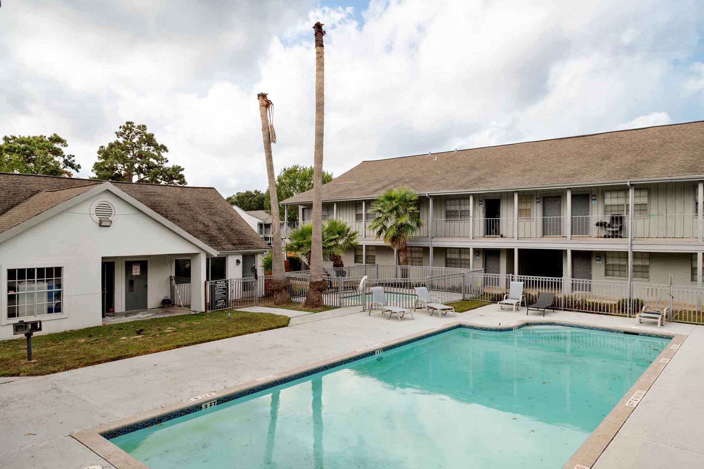 Swimming pool surrounded by a concrete deck, flanked by white buildings and palm trees. In the background, a two-story building features balconies. A couple of palm trees have been partially cut down. The sky is cloudy, giving a serene atmosphere to the scene.