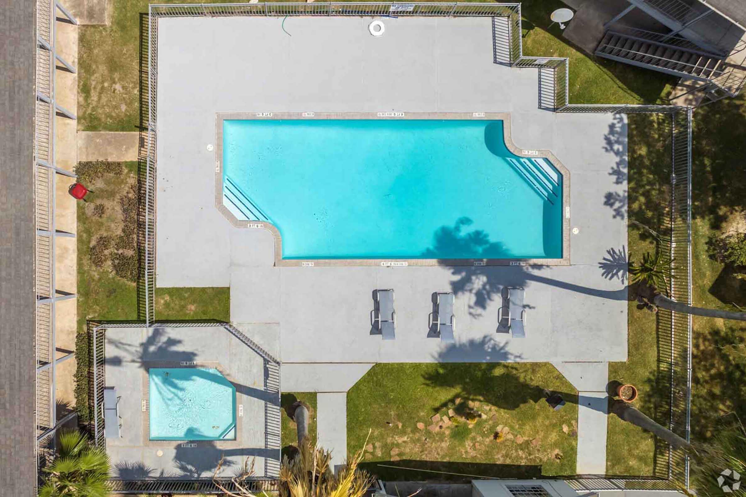 Aerial view of a swimming pool area featuring a large rectangular pool with clear blue water, surrounded by a gray concrete deck. There are four lounge chairs around the pool and a smaller hot tub adjacent to it, all enclosed by a fence. Lush greenery and palm trees are visible nearby.