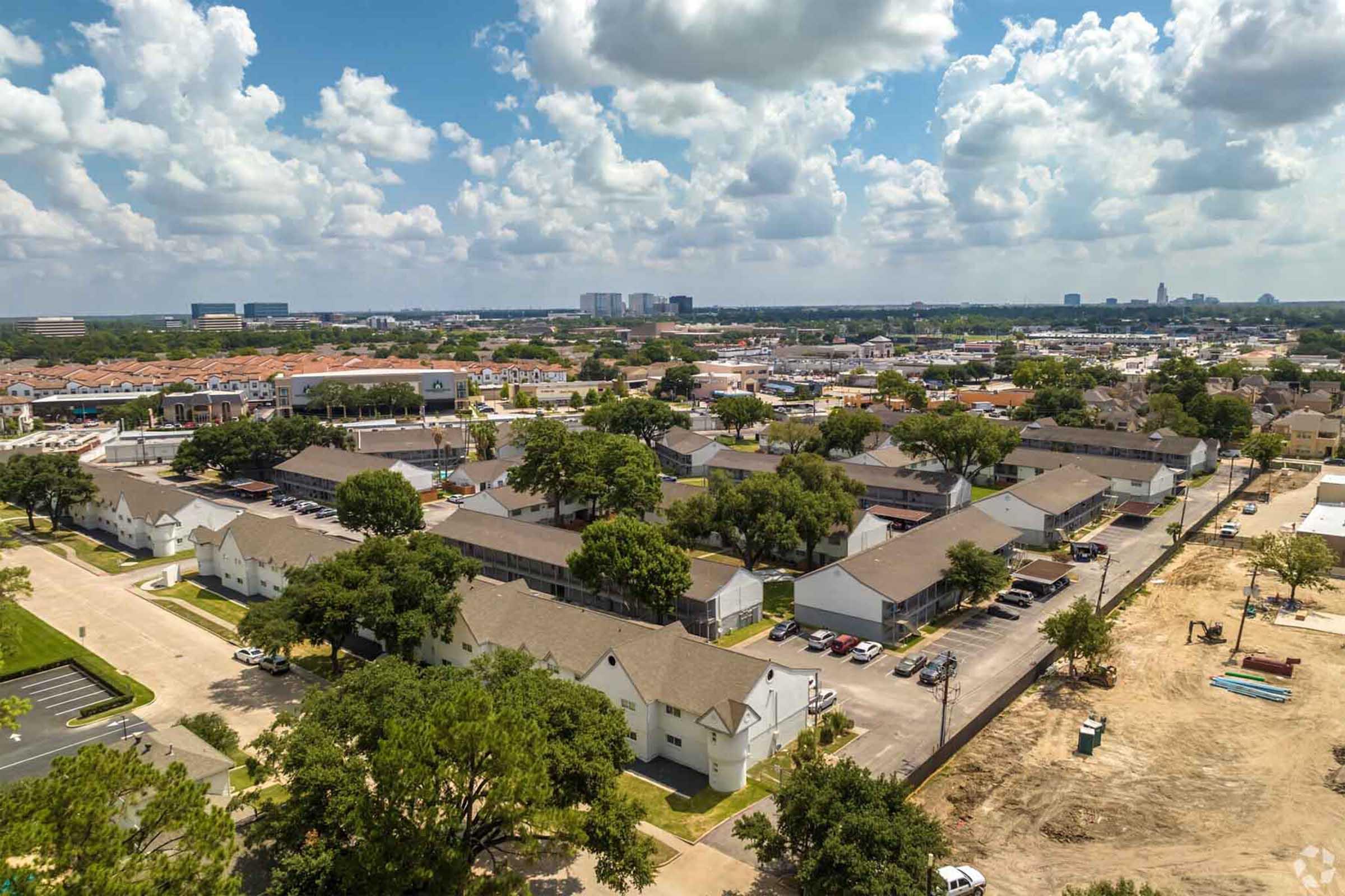 Aerial view of a suburban neighborhood featuring clusters of low-rise apartment buildings, scattered trees, and parking areas. In the background, there are commercial buildings and a skyline under a partly cloudy sky. A construction site is visible in the lower right corner.