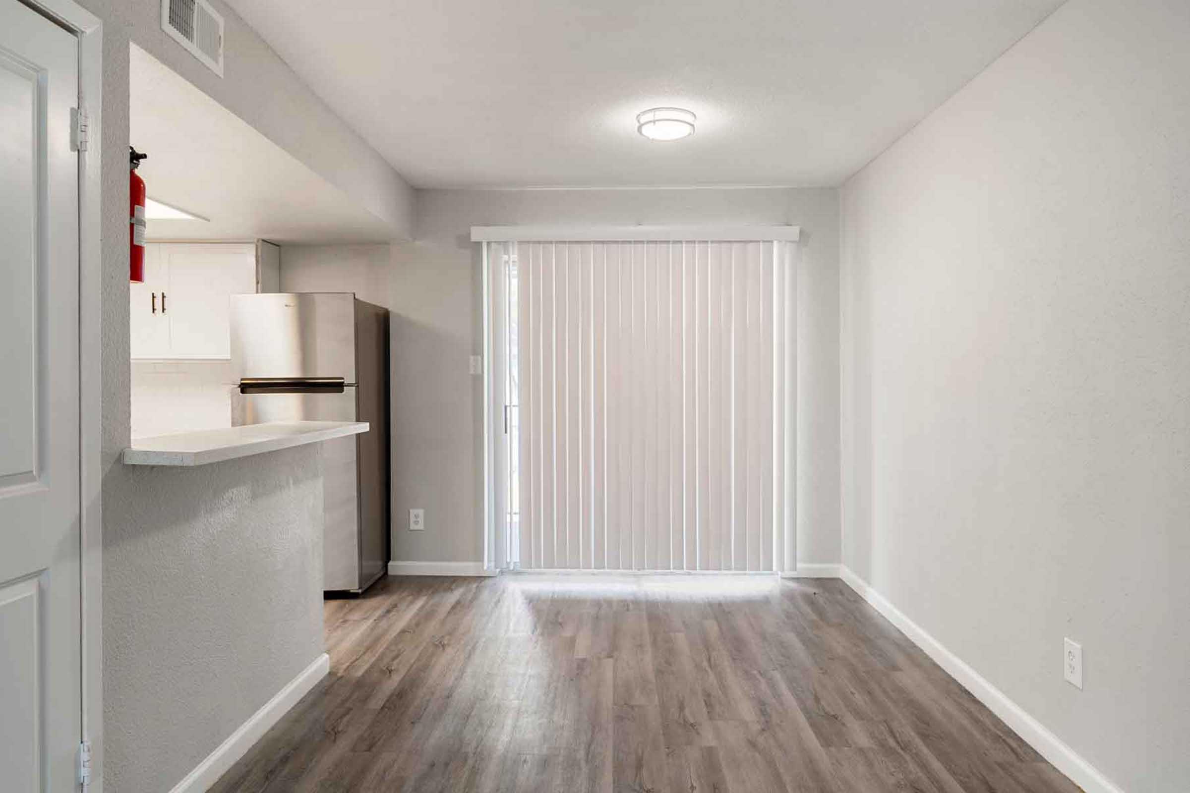 A view of a modern, minimalistic apartment interior featuring a light-colored wall, wooden flooring, and a sliding glass door with vertical blinds. To the left, there's a small kitchen area with a refrigerator, and the space is well-lit with a ceiling light, creating an open, inviting atmosphere.