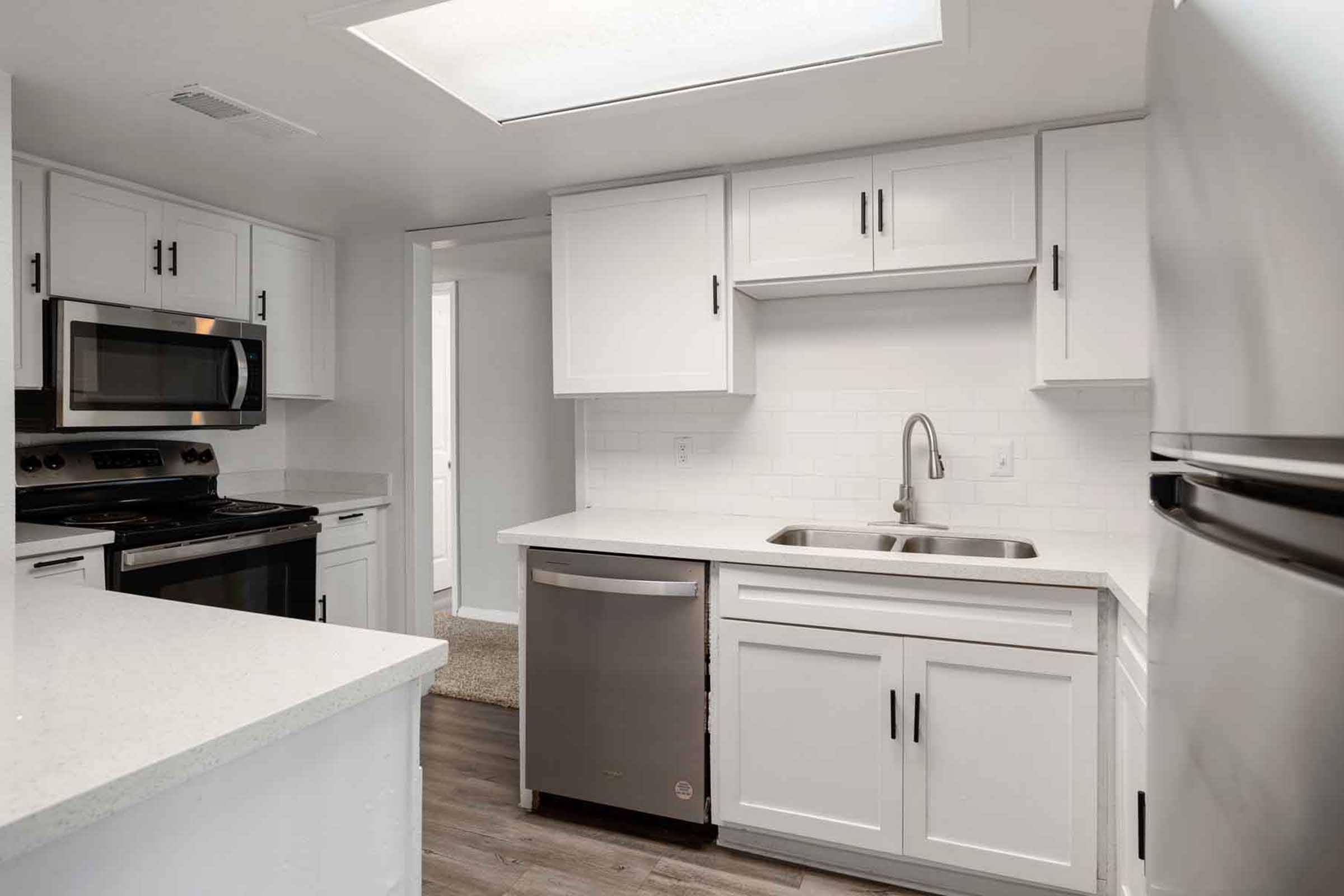 Modern kitchen featuring white cabinetry, stainless steel appliances including an oven, microwave, and refrigerator, a single bowl sink, and a large countertop with a clean design. Natural light comes in from a skylight, enhancing the bright and airy feel of the space. The flooring is a warm, muted tone.