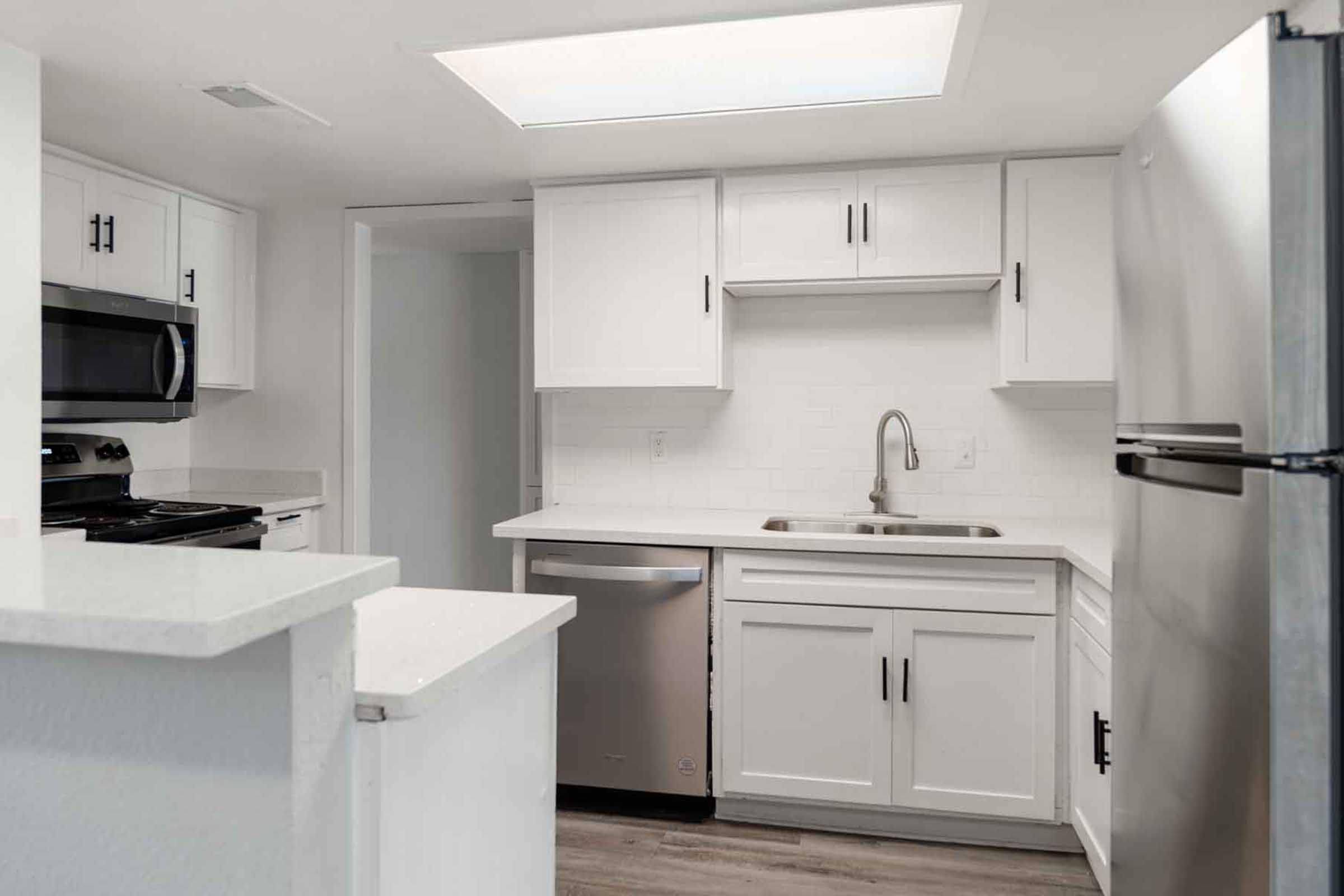 A modern kitchen featuring white cabinetry, a stainless steel refrigerator, a sink, and a stove. The countertops are light-colored, and there is a prominent skylight above for natural lighting. The flooring is a light wood laminate, and the space appears clean and organized.