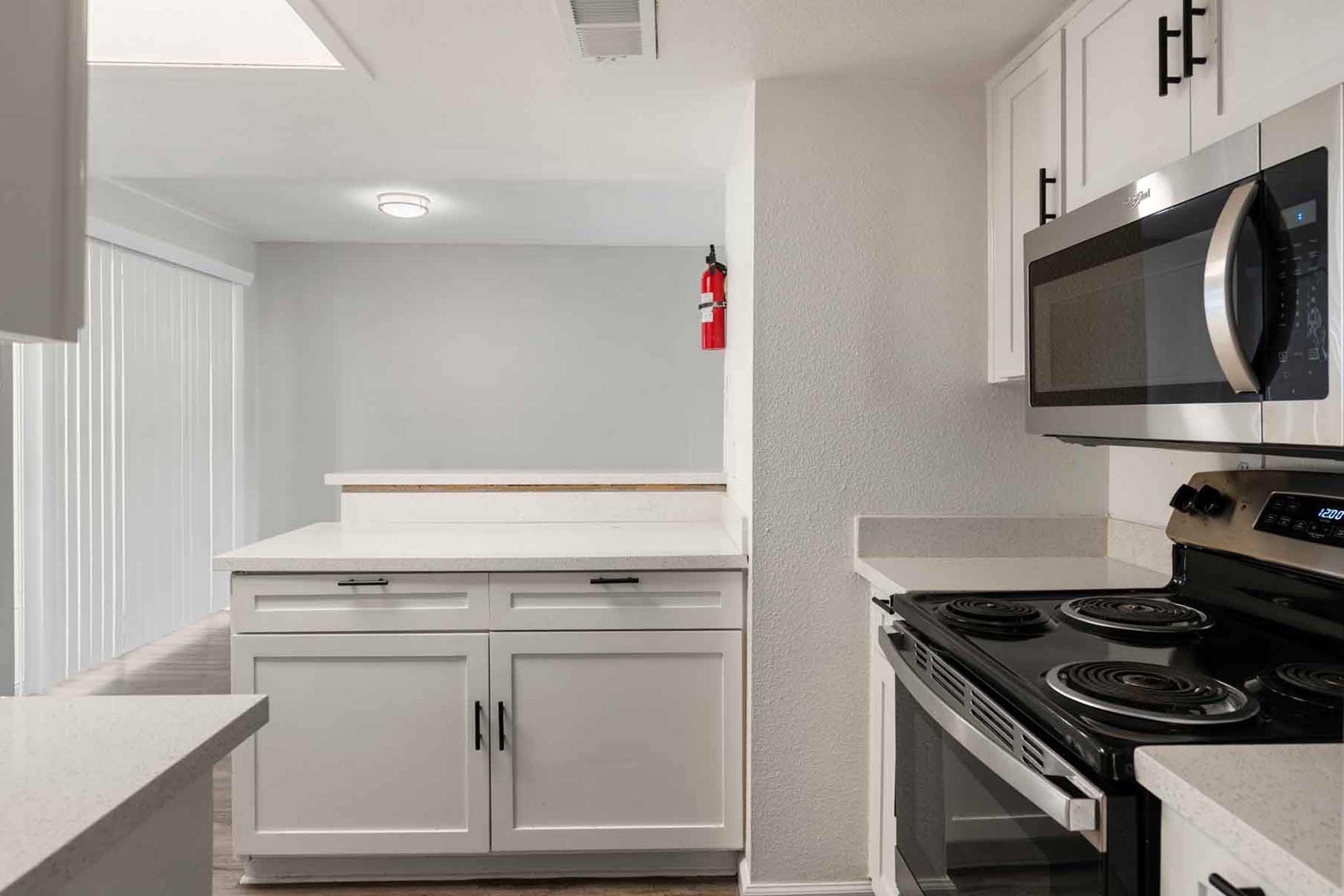 Modern kitchen featuring white cabinets and countertops, a microwave, and a black stove. The space is well-lit, with a glimpse of a gray wall in the background and a fire extinguisher mounted on the wall. Smooth flooring complements the contemporary design.