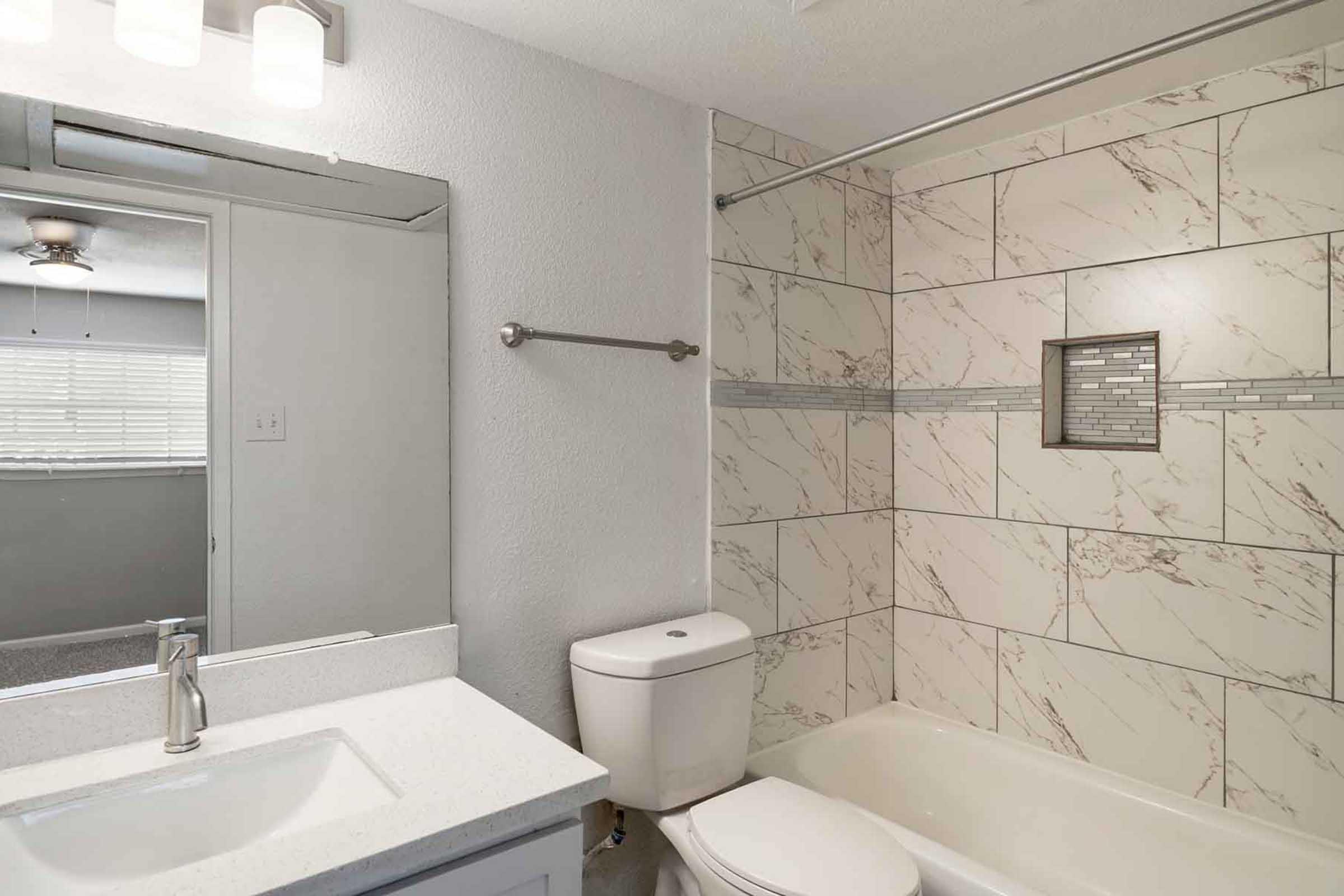 A modern bathroom featuring a white vanity with a sink, a toilet, and a bathtub with a shower. The walls are tiled with a light marbled pattern, and there's a small decorative inset. A mirror hangs above the sink, and a window provides natural light.