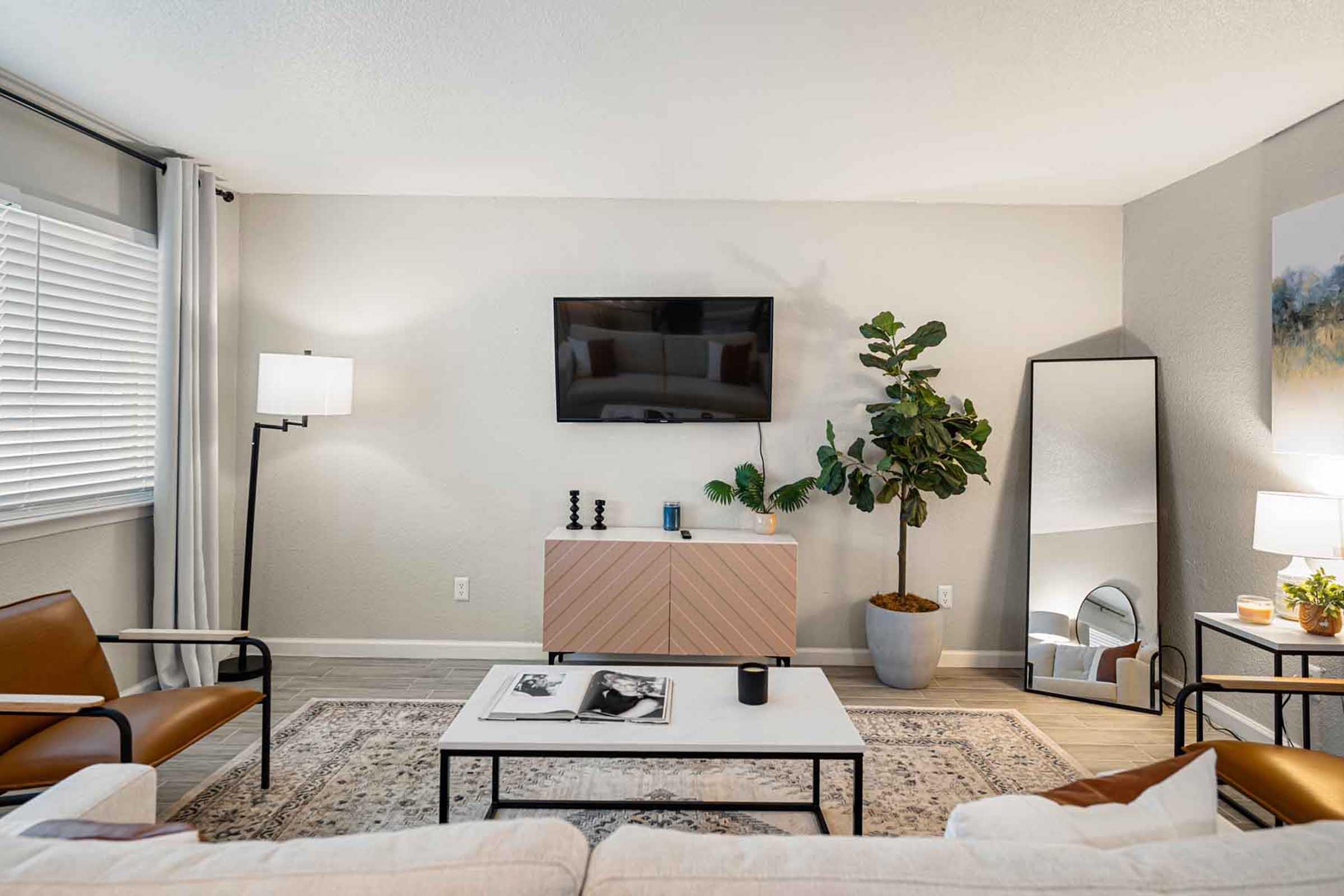 A modern living room featuring a beige sofa, two chic chairs, a wooden side table, and a large flat-screen TV mounted on the wall. A tall indoor plant adds greenery, while a round mirror and stylish decor enhance the aesthetic. Natural light filters through light curtains, creating a welcoming atmosphere.