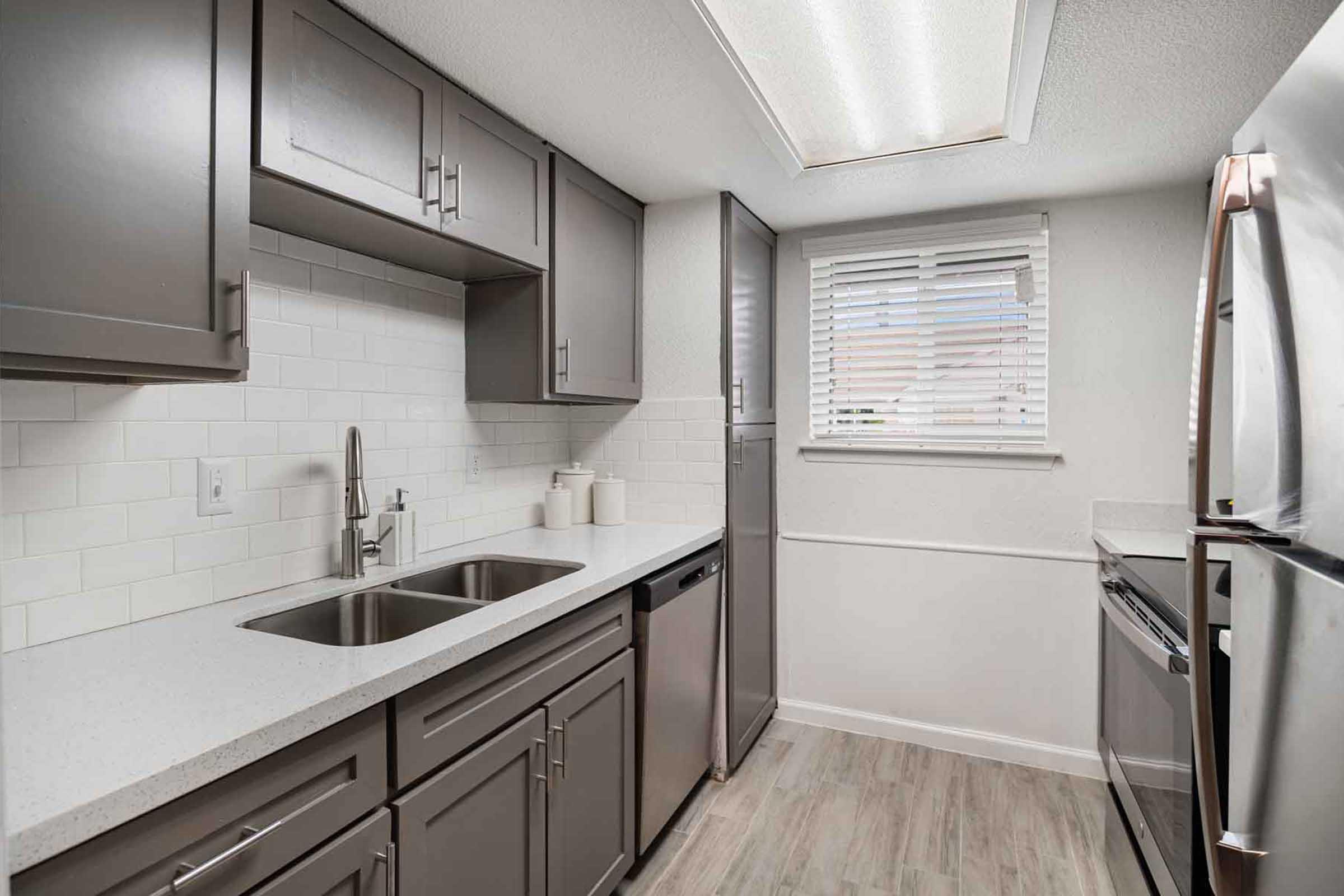 Modern kitchen featuring gray cabinetry, a double sink, stainless steel appliances, and white subway tile backsplash. The space has natural light from a window with blinds and a skylight above. The floor has light wood-like tiles, creating a bright and open atmosphere.