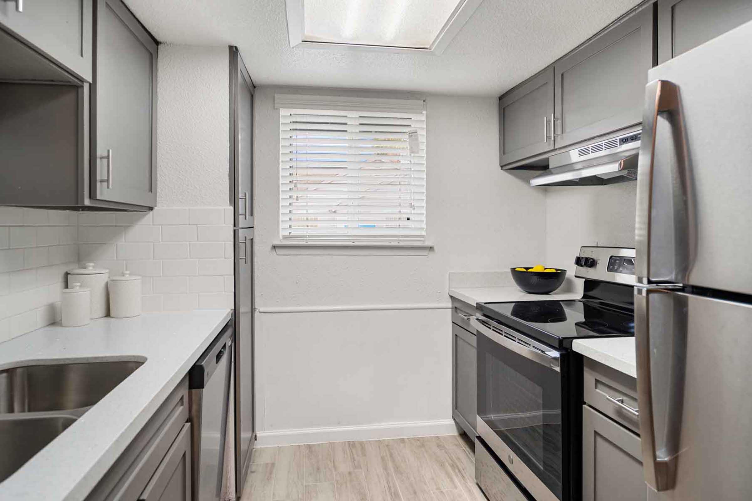 Modern kitchen with gray cabinets and white countertops. It features stainless steel appliances, including an oven and fridge, a window with blinds, and a bowl of yellow fruit on the countertop. The flooring is light, adding a spacious feel to the area.